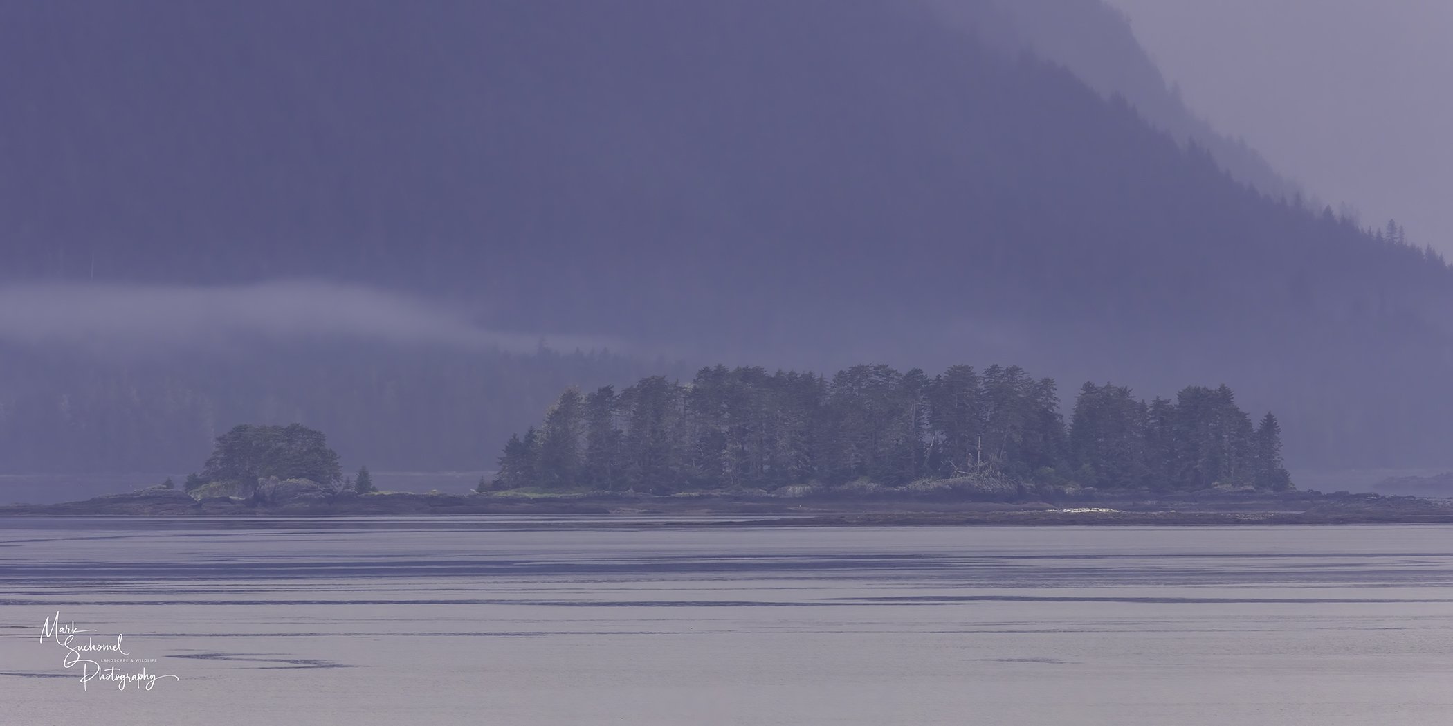 A tranquil seascape showing multiple tree-covered islands and distant mountains, with mist or fog over the water.