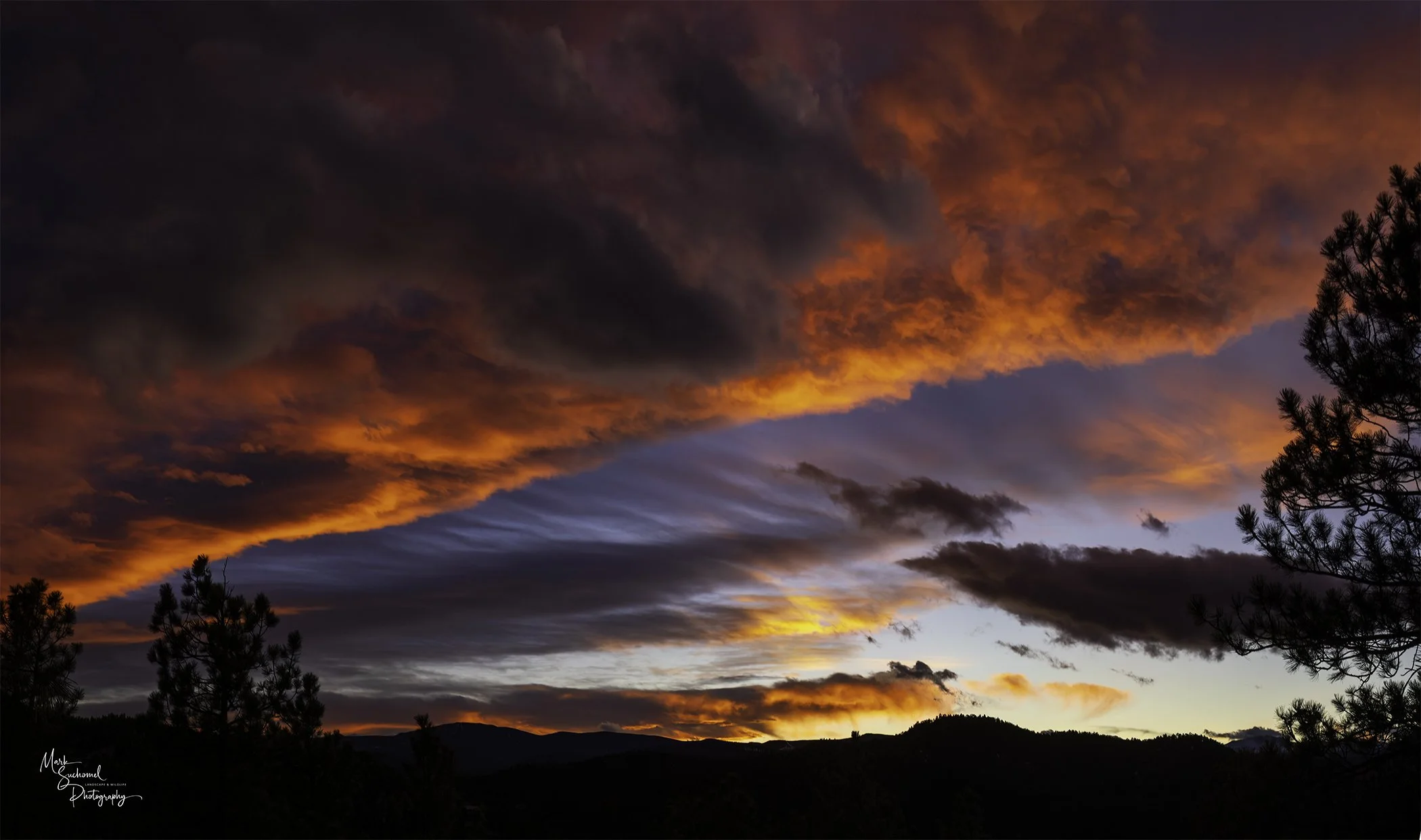 Colorful sunset sky with dark clouds, silhouettes of trees and mountains in the foreground.
