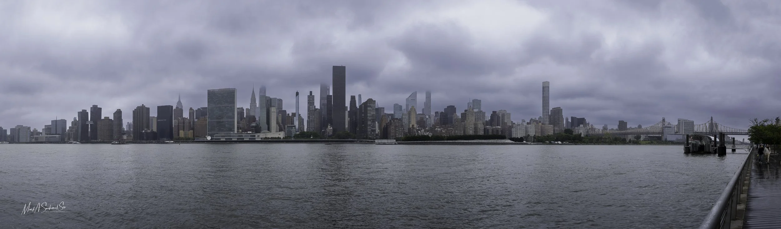 Overcast view of Manhattan skyline from across the river, with tall buildings, some partially obscured by clouds, and a nearby pier on the right.