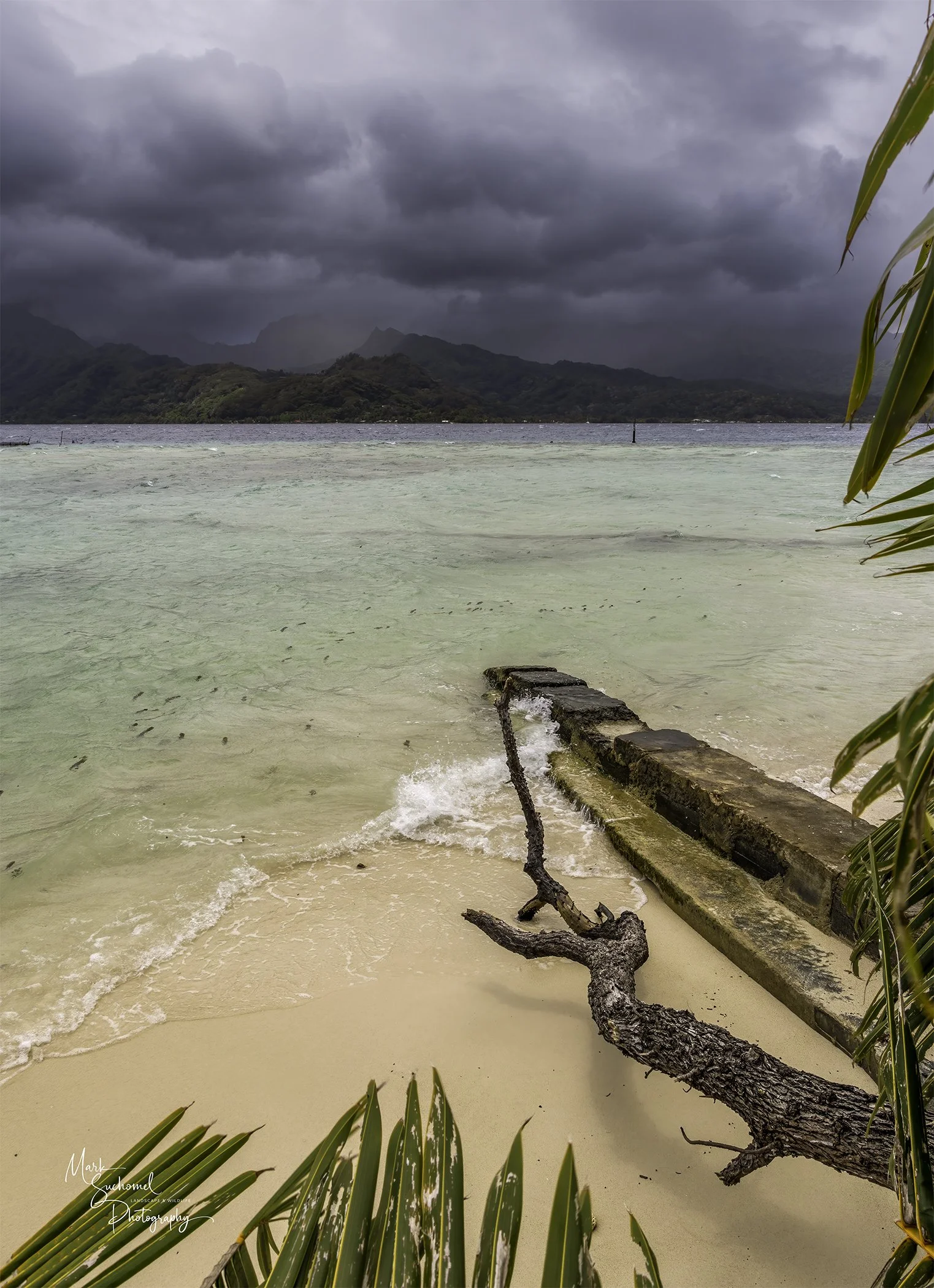 Stormy weather over a beach with a cloudy sky, green mountains in the distance, and a piece of driftwood on the sandy shore near a concrete structure.