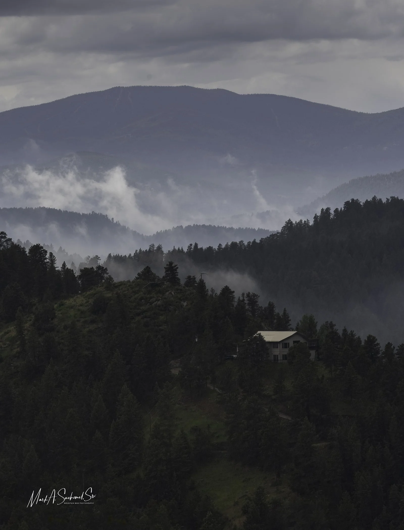 Mountain view with layers of fog and trees, a house visible on a hillside, under cloudy sky.
