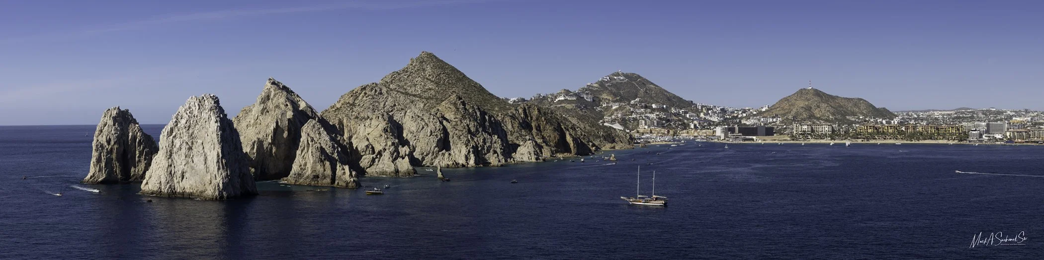 Scenic view of rocky cliffs along the coast with boats sailing on the water, a cityscape, and mountains in the background under a clear blue sky.