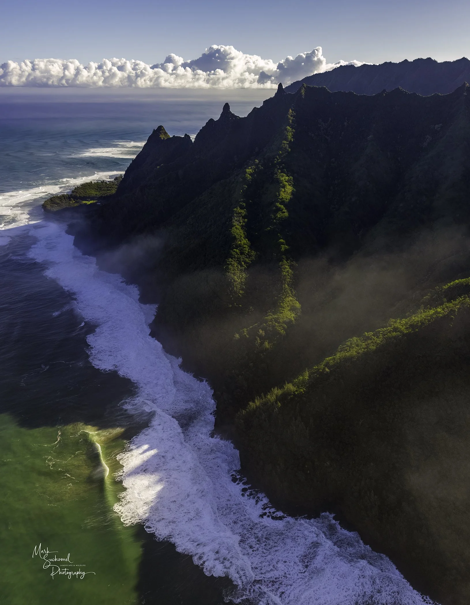 Aerial view of rugged green mountains along the coastline with the ocean, waves crashing on the shore, and clouds in the sky.