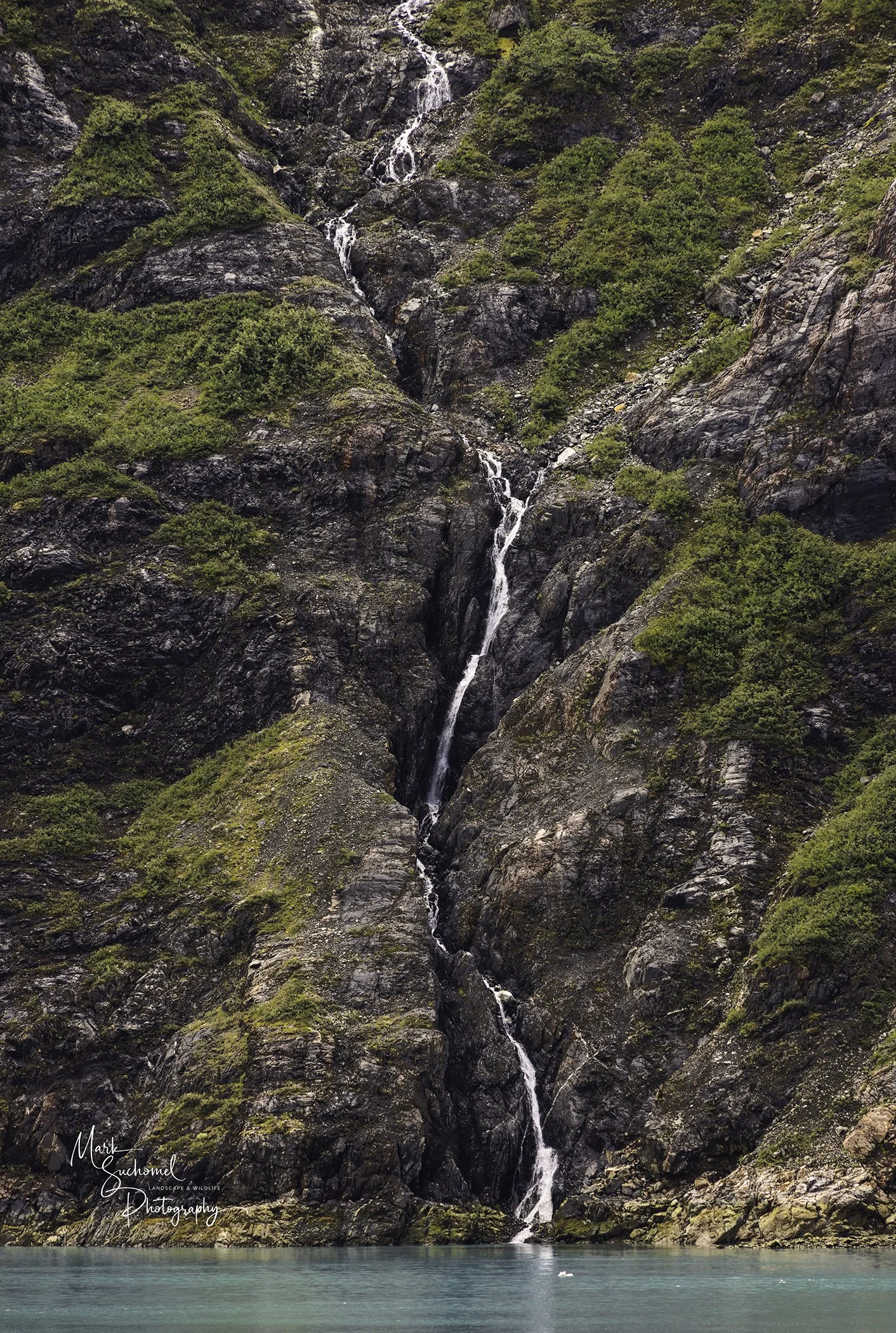 A tall, narrow waterfall cascading down dark, rugged rocks surrounded by green moss and bushes, flowing into a turquoise body of water at the bottom.