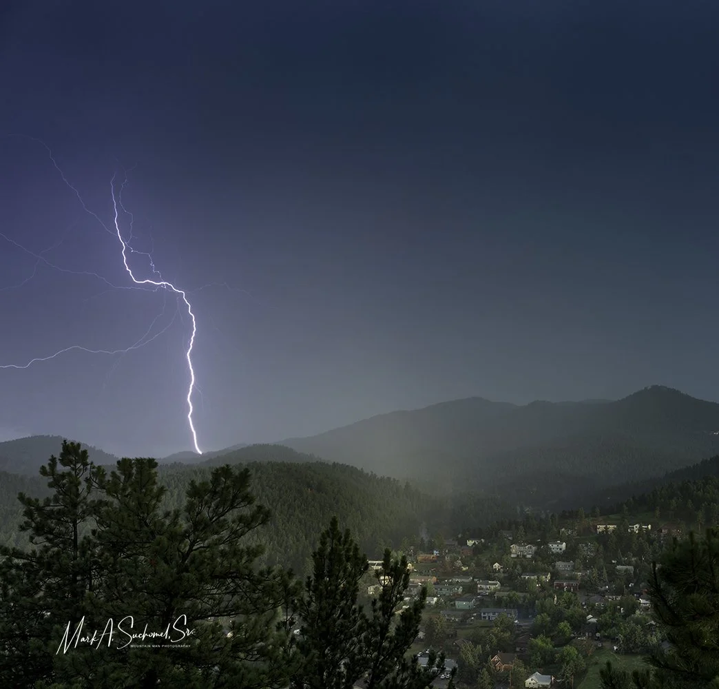 Lightning strikes over a mountainous landscape with a small town below, dark sky in the background.