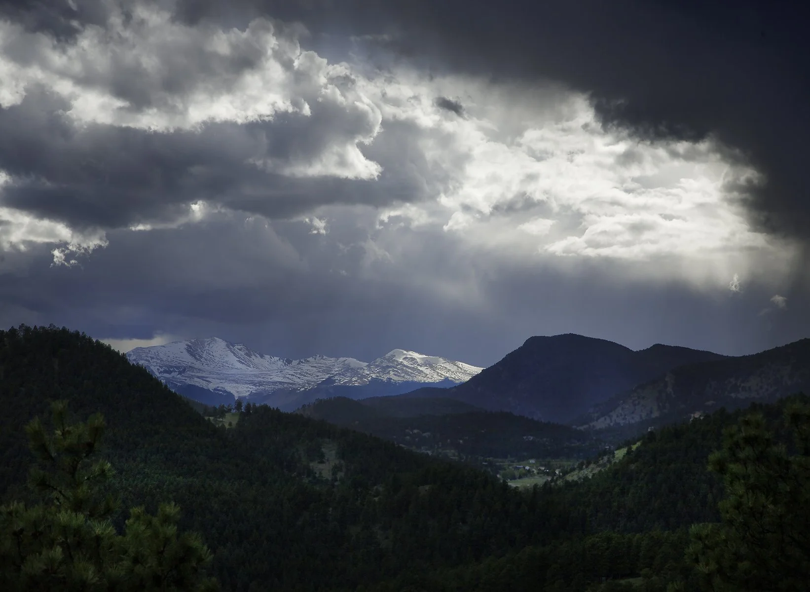 Mountains with snow-capped peaks under a cloudy sky, with a forested hillside in the foreground.
