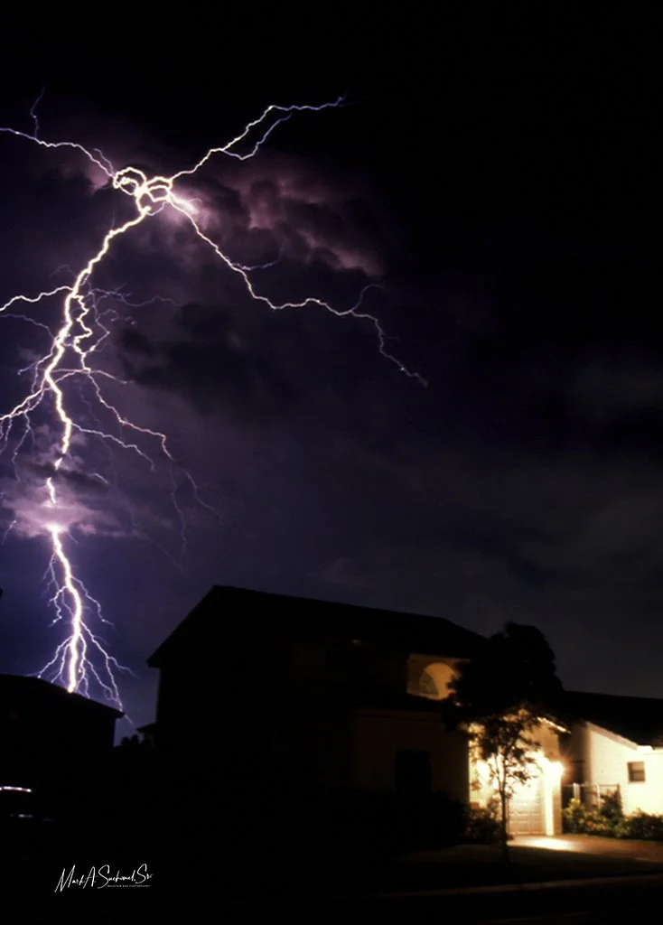 Lightning striking in a dark night sky above houses with illuminated windows and a tree in front.