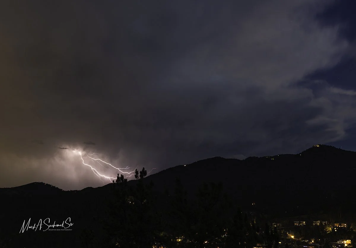 Nighttime view of lightning strikes in the sky over a mountain range, with dark clouds and illuminated city lights below.