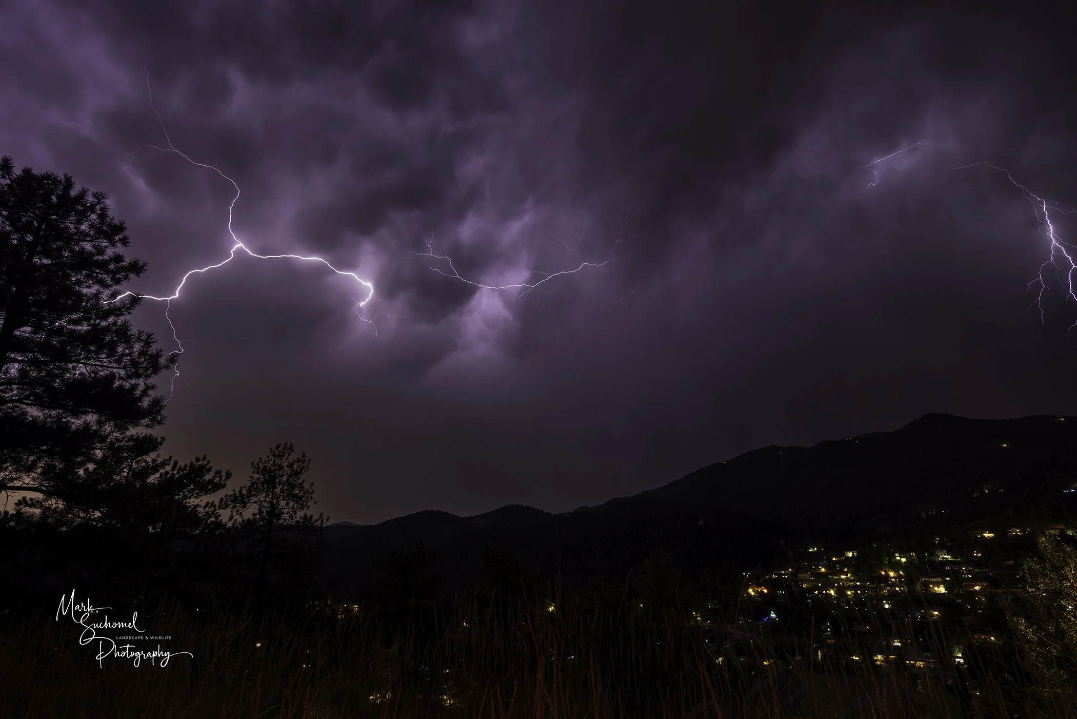 Lightning strikes illuminate a dark stormy sky over a mountainous landscape with a small town at the base. Tall trees are silhouetted in the foreground.