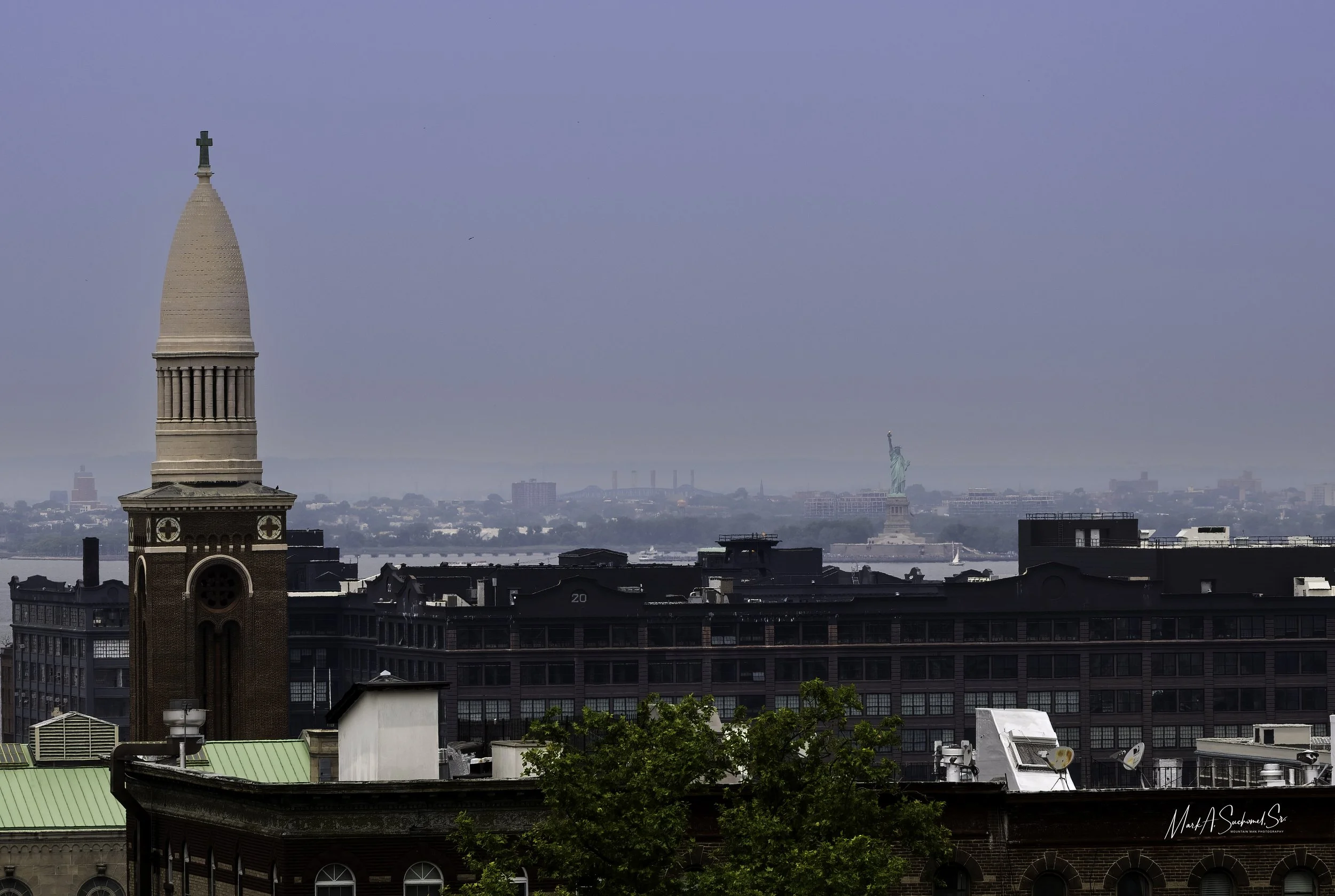 A city skyline with a church steeple in the foreground, the Statue of Liberty in the distance on the water, and a clear blue sky.