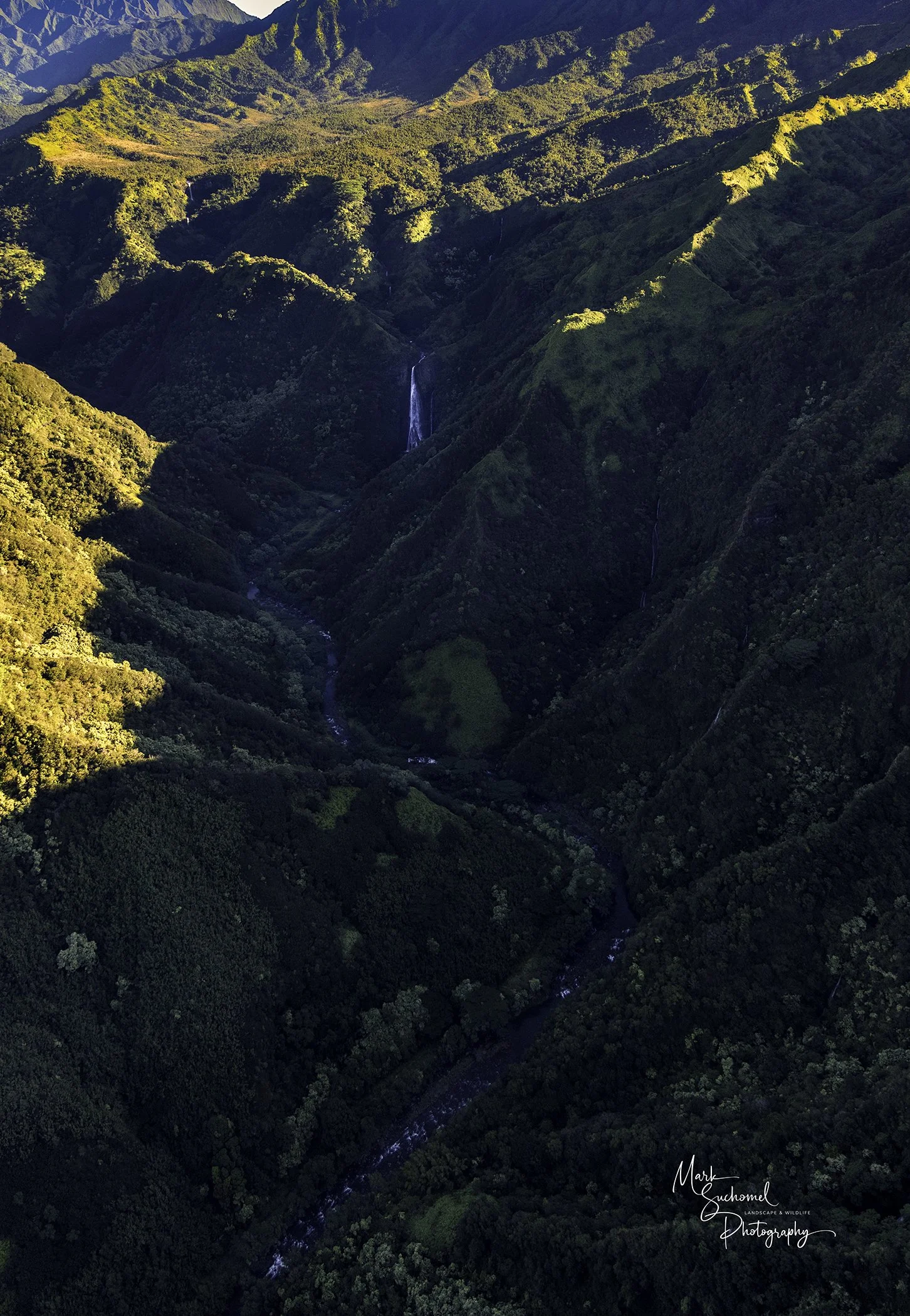 Aerial view of lush, green mountains with a small waterfall and a narrow river running through the valley.