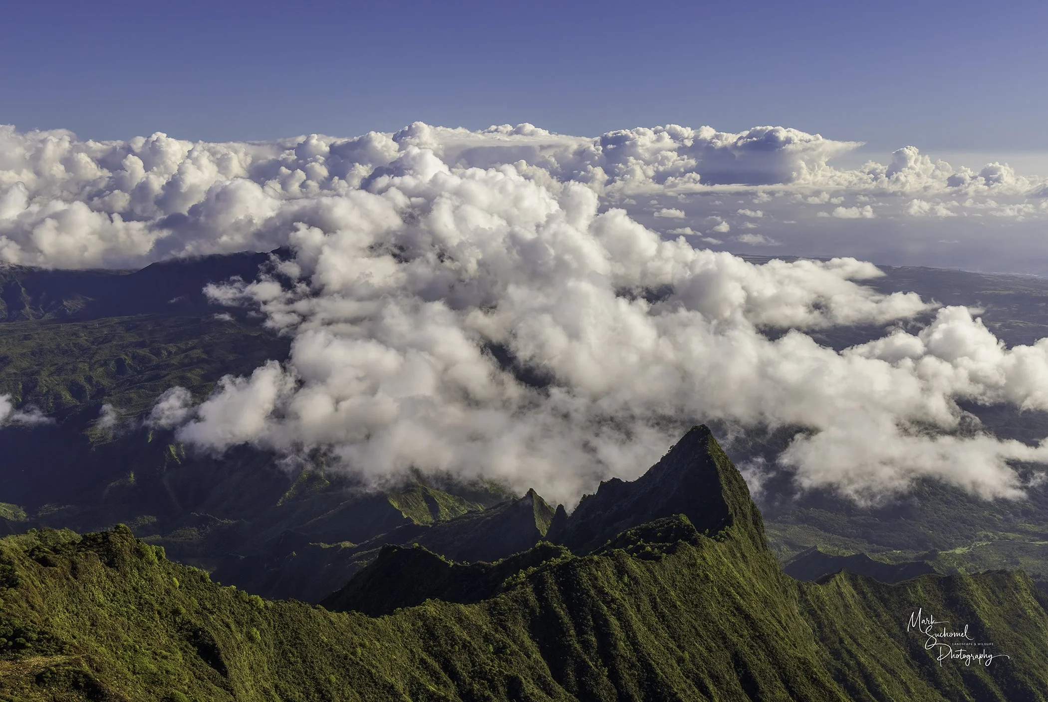 A mountain ridge covered in green vegetation with sharp peaks, surrounded by clouds and a blue sky.