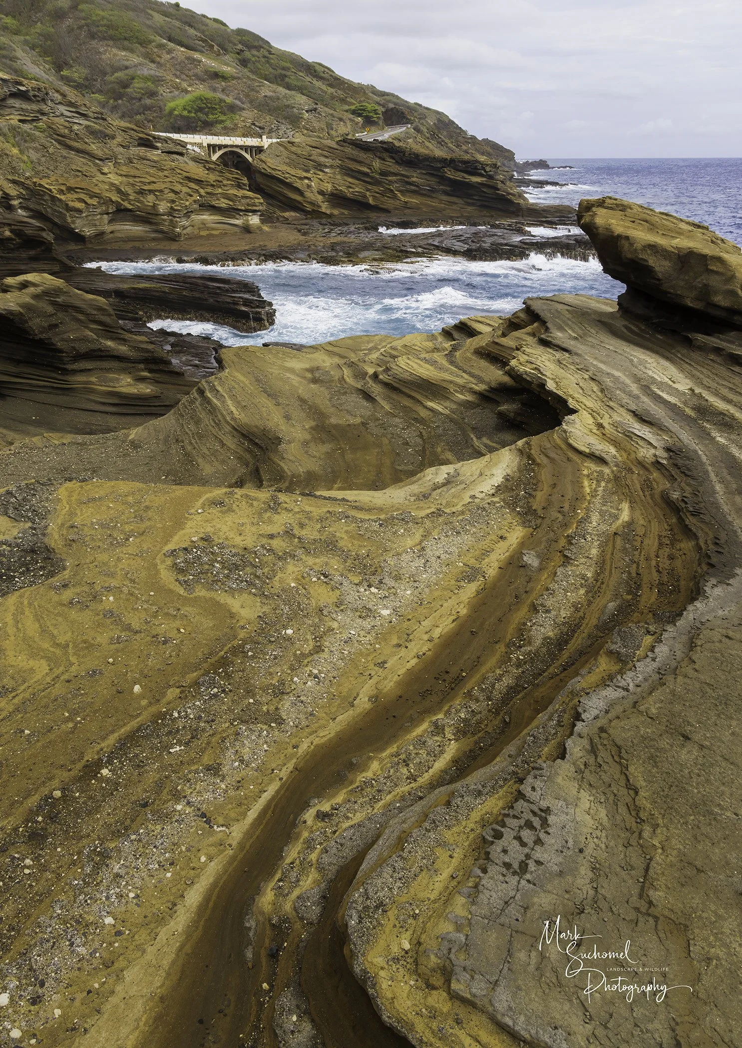 Colorful layered rock formations along a rugged coastline with waves crashing against the rocks, and a hillside with sparse vegetation and a curved road in the background.