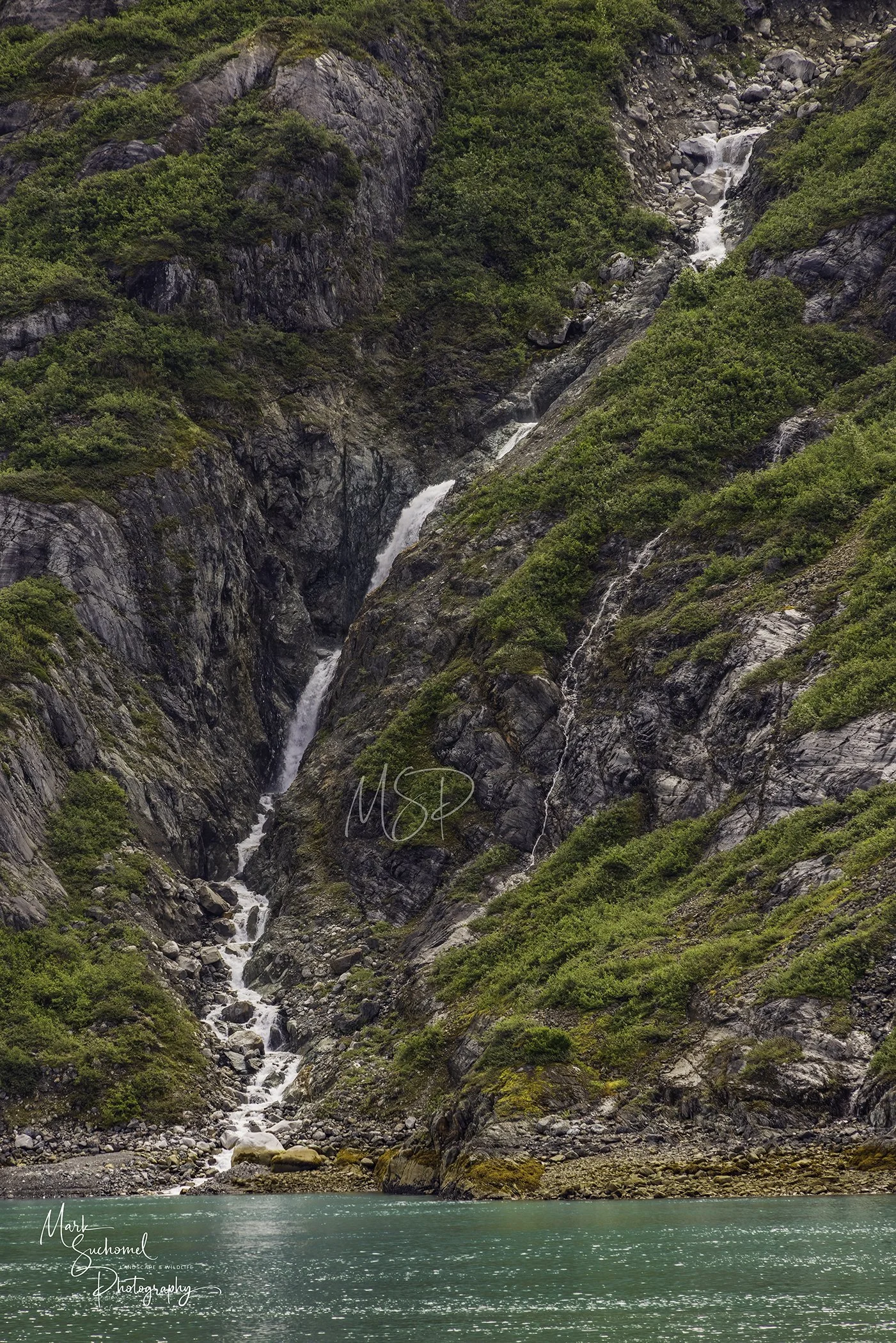 A mountain with green vegetation and rocky cliffs, with a small waterfall flowing down into a turquoise body of water at the bottom.