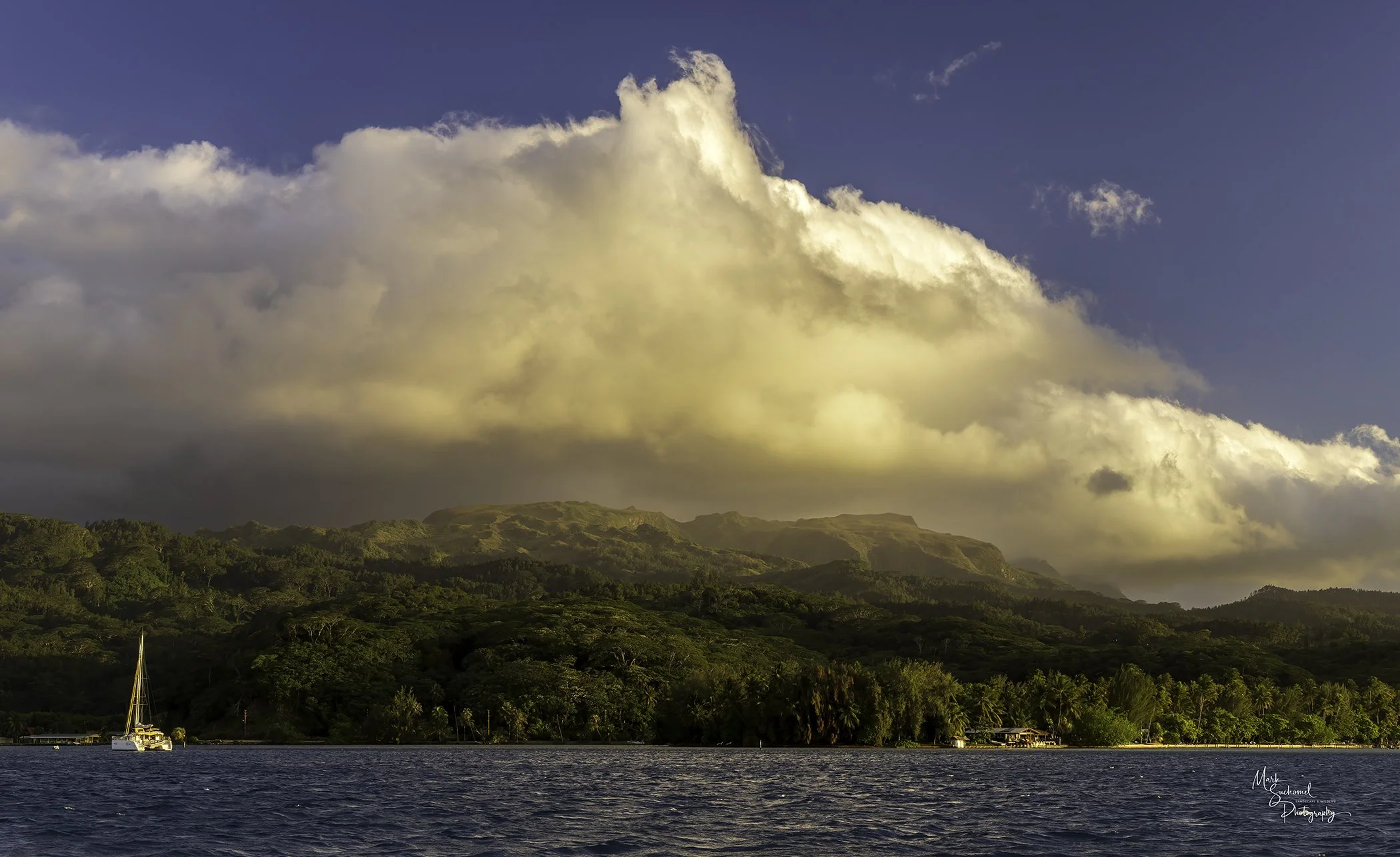 View of a boat on calm water with lush green hills and mountains in the background, under a partly cloudy sky during sunset.