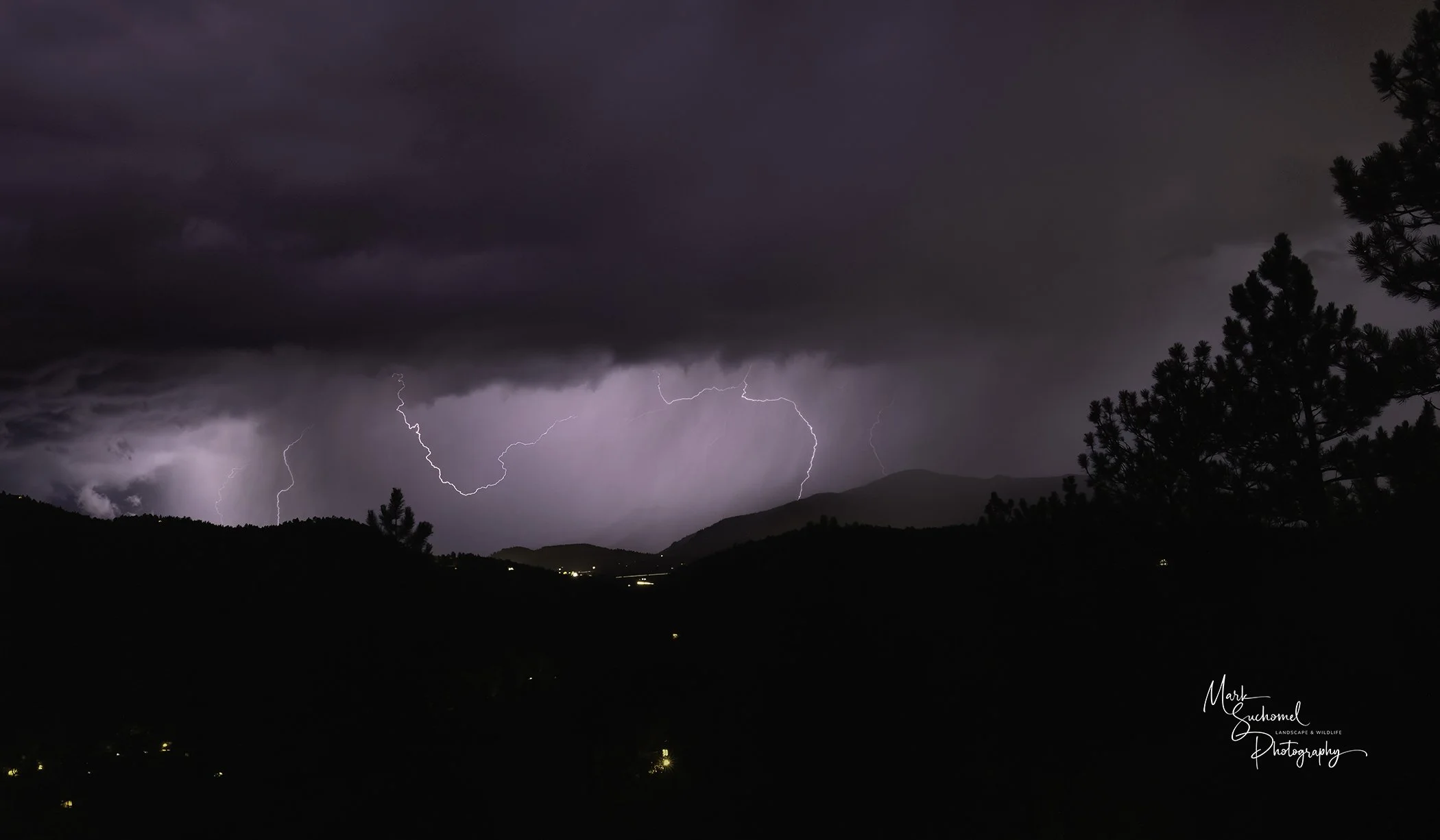 Nighttime landscape with dark clouds and multiple lightning bolts striking through the sky over mountains, with a few scattered lights in the valley. Forested hills are silhouetted in the foreground.