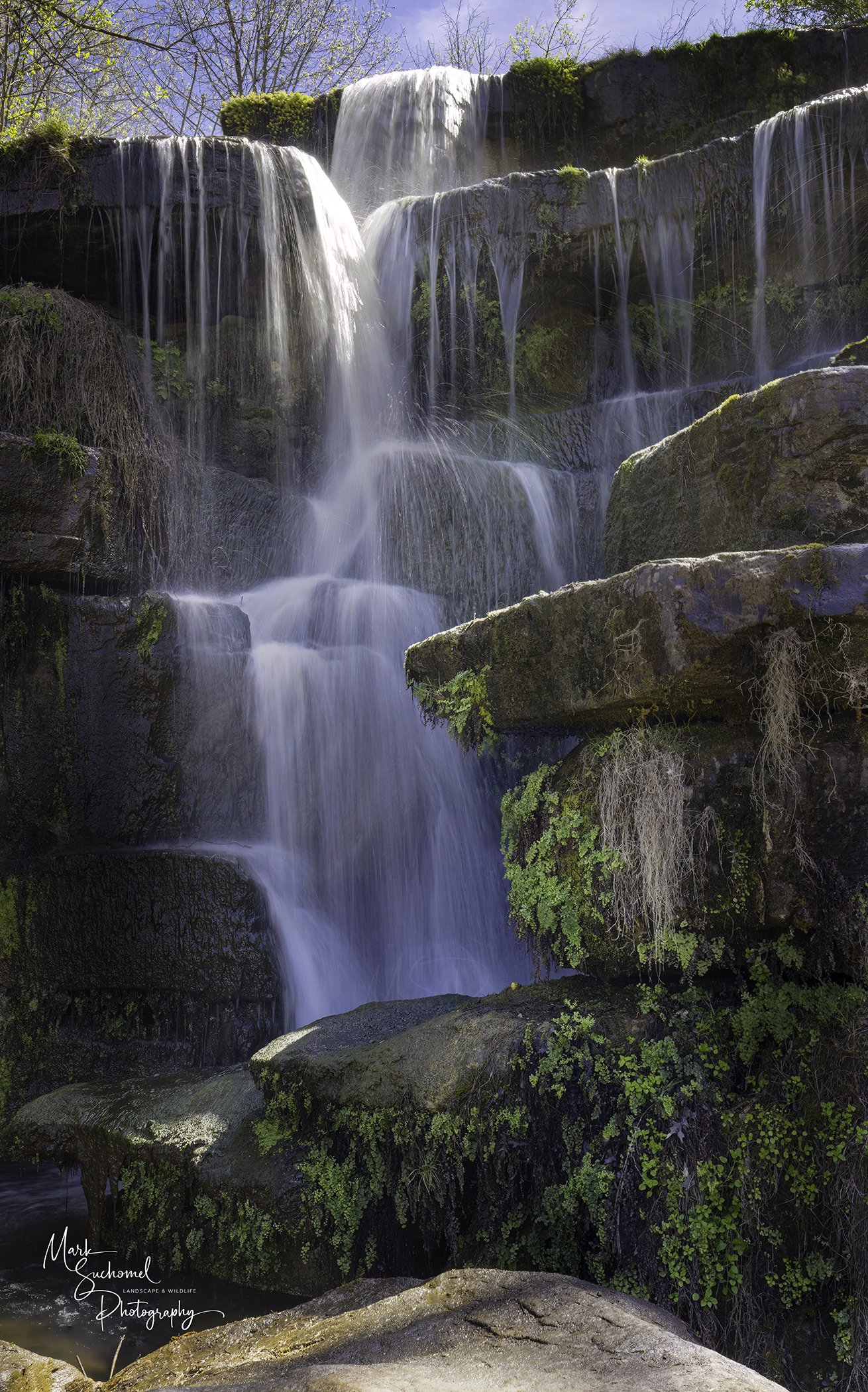 A multi-tiered waterfall flowing over rocks with lush green moss and plants, under a clear blue sky.