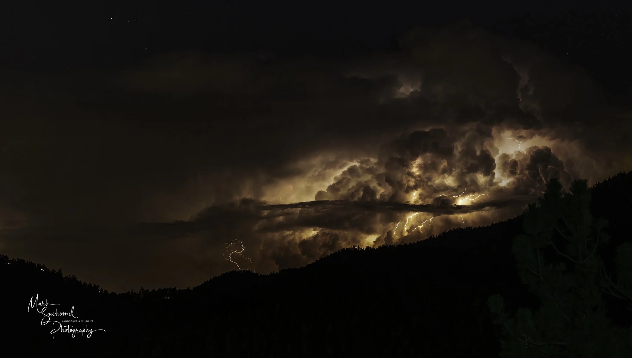 Nighttime thunderstorm with lightning illuminating large, dark storm clouds over a mountainous landscape, with trees silhouetted in the foreground.