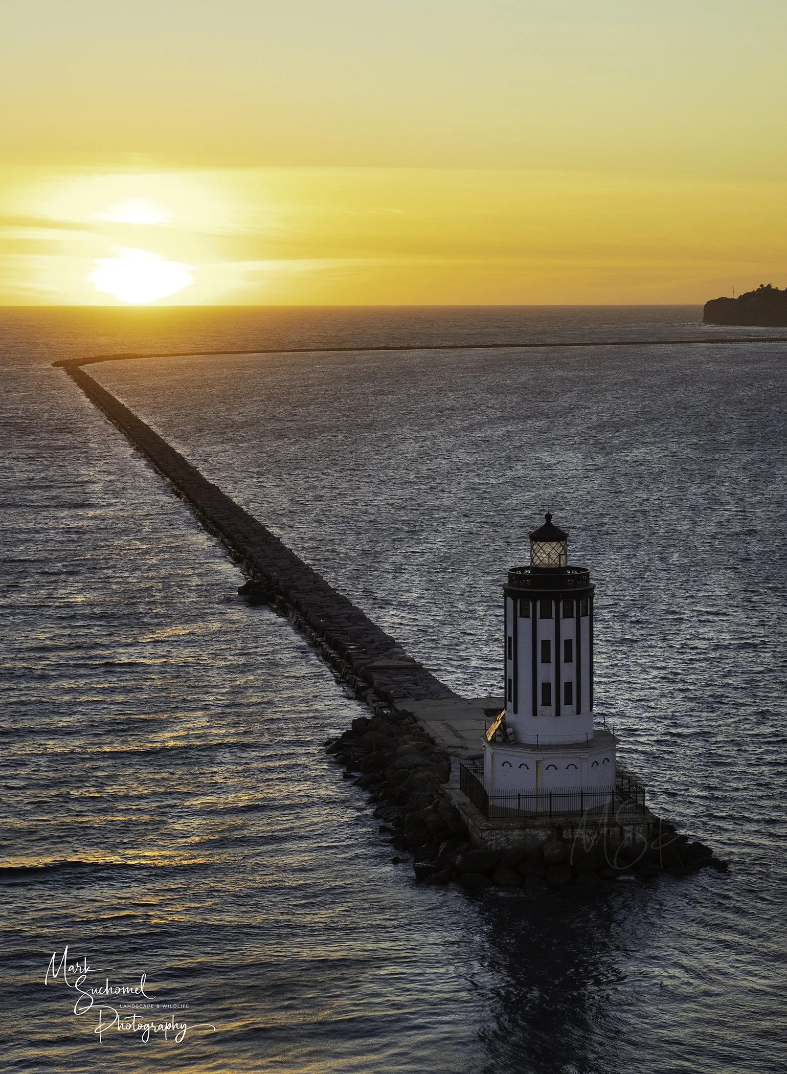A lighthouse on a rocky pier extends into the ocean at sunset, with the sun near the horizon and the sky painted in orange and yellow hues.