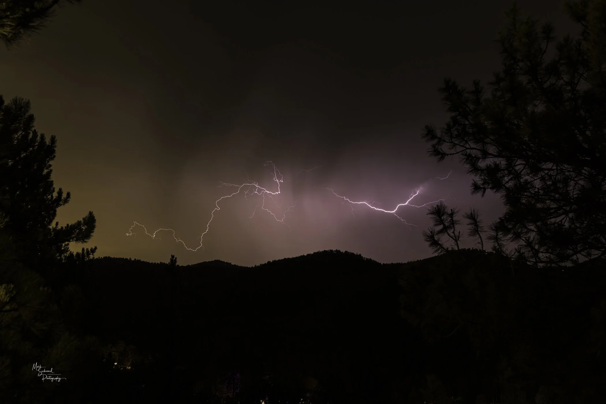 Lightning strikes across a cloudy night sky over silhouetted mountain landscape with trees on the foreground.