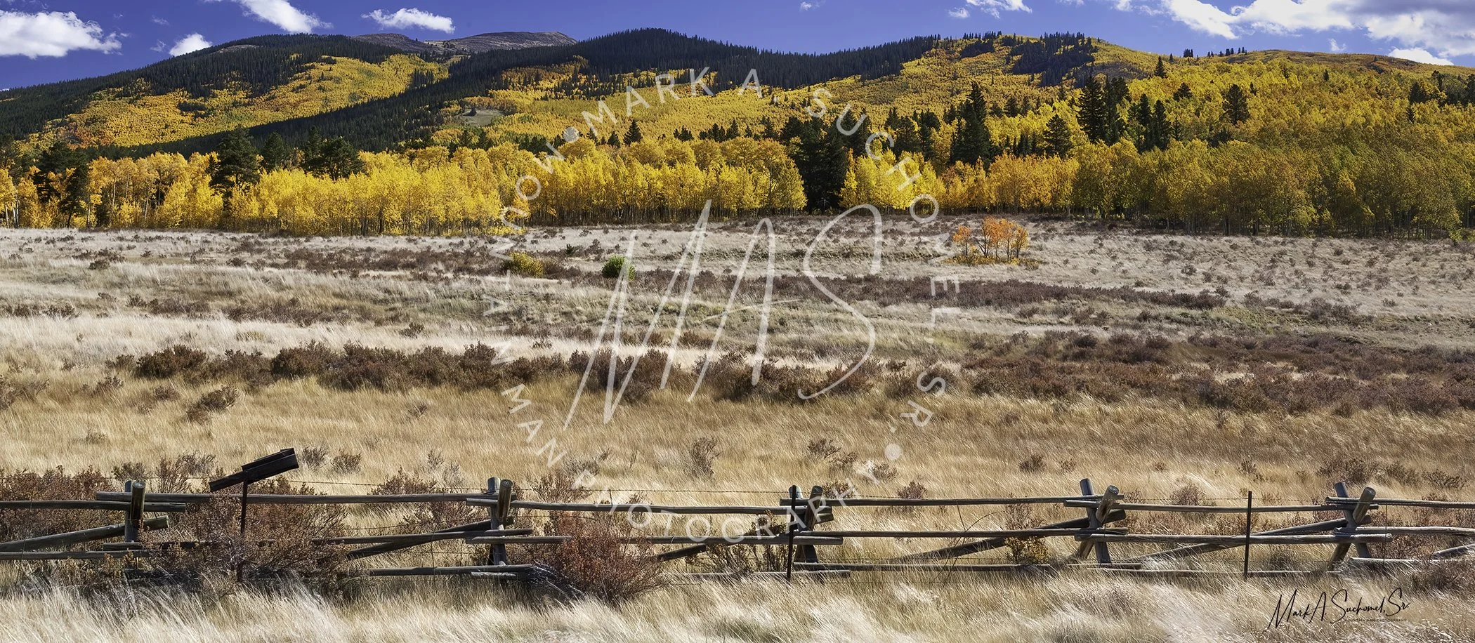A scenic view of a mountain landscape in autumn, featuring colorful fall foliage on the trees, a yellow grassy field in the foreground, and a wooden fence in the front. The sky is partly cloudy with patches of blue.