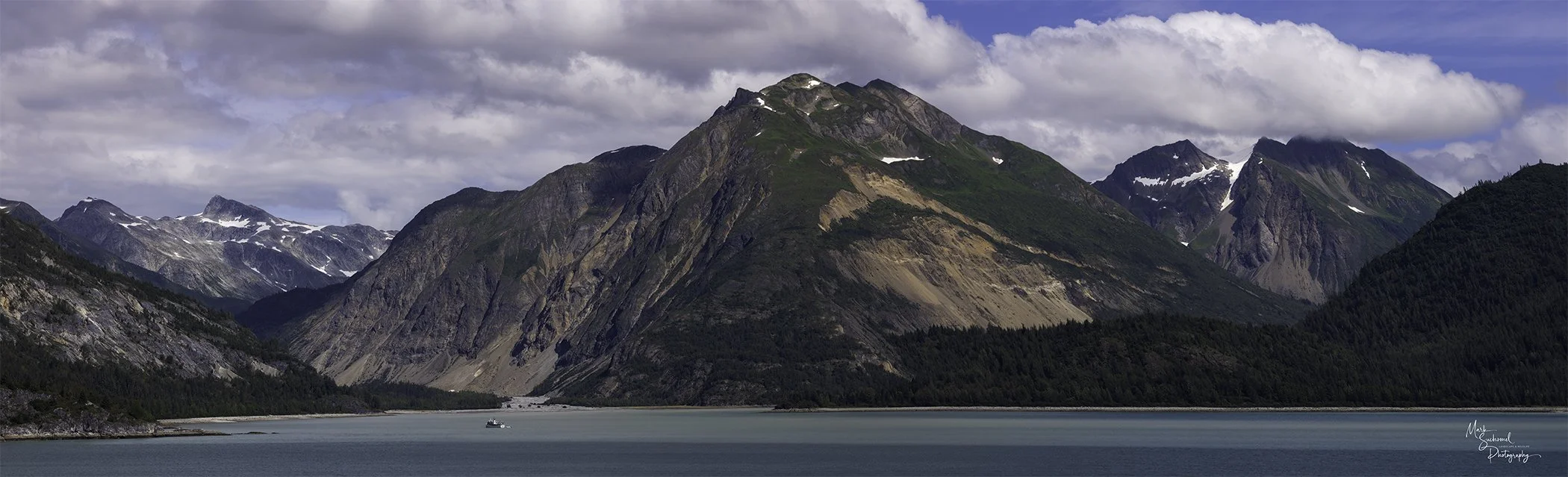 A scenic view of a large body of water with mountains in the background, some with patches of snow on their peaks and slopes. The sky is partly cloudy.
