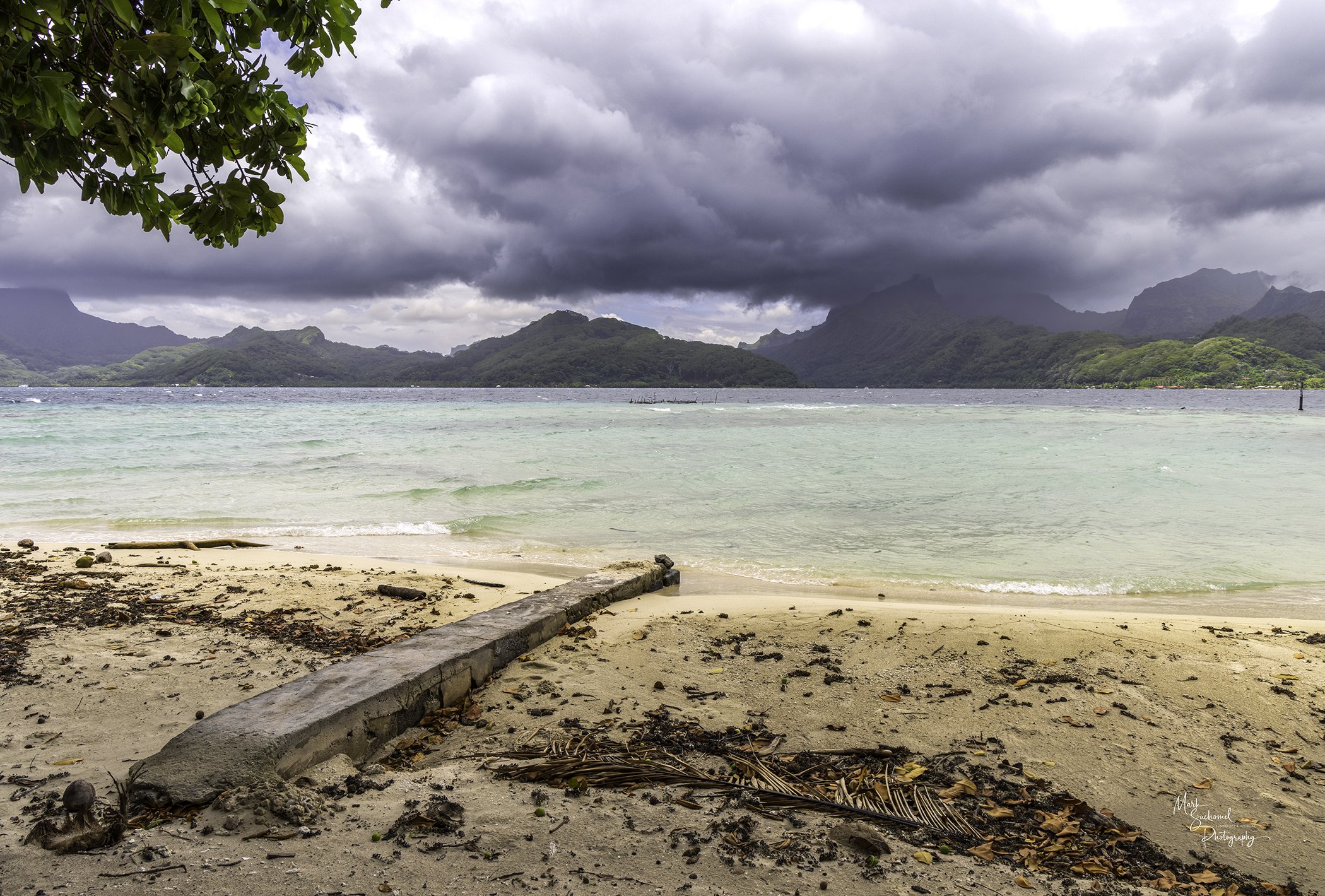 A beach scene with cloudy skies, green mountainous landscape in the background, sandy shore with debris, and a leaning wooden beam in the foreground.