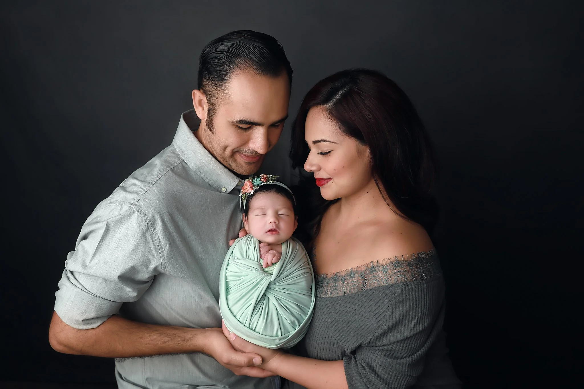 A loving family from Newport Beach, California, poses for a professional newborn portrait. The mother and father gaze adoringly at their sleeping baby, who is swaddled in a soft mint green wrap with a delicate floral headband. The intimate and heartw