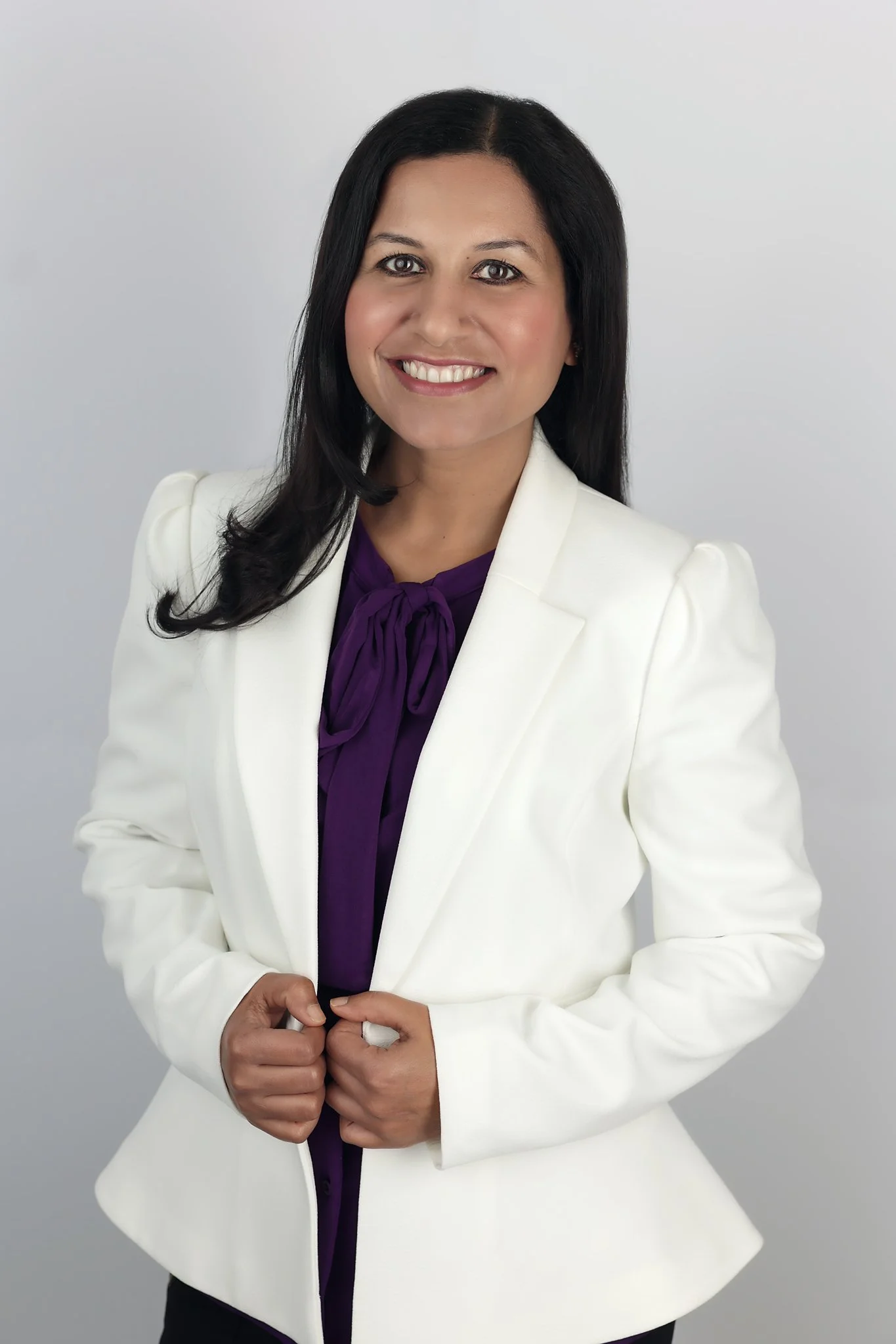 A professional headshot of a realtor from Irvine, California, wearing a white blazer over a purple blouse. She smiles confidently against a clean, neutral background, showcasing a polished and approachable presence.