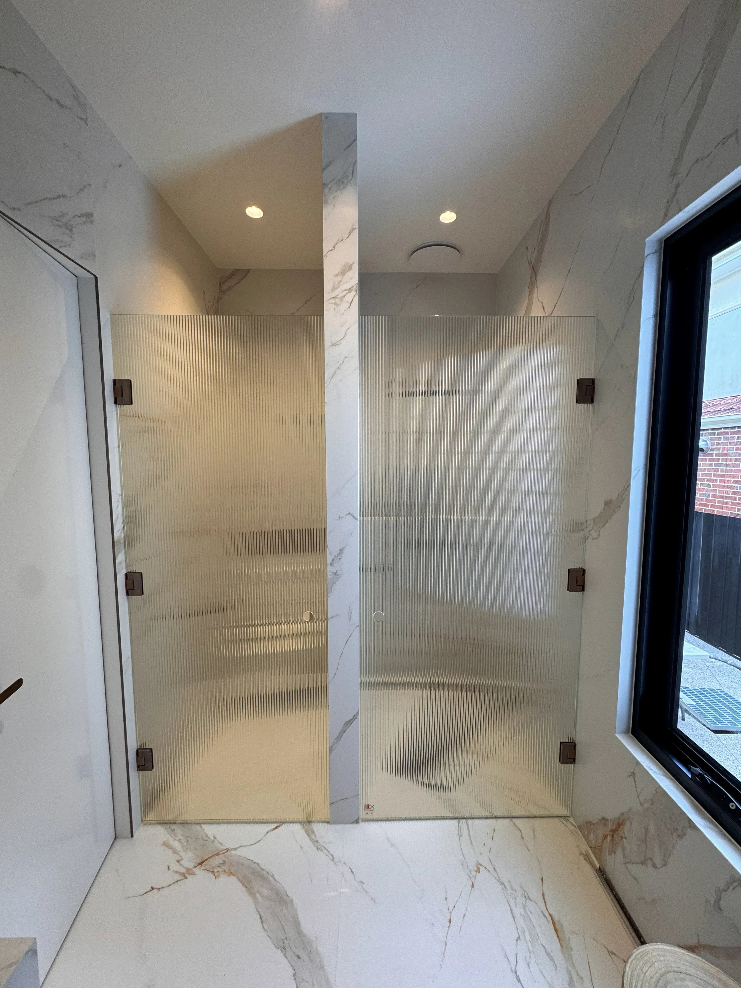Interior view of a modern bathroom with frosted glass shower doors, marble walls, and a window with black framing.