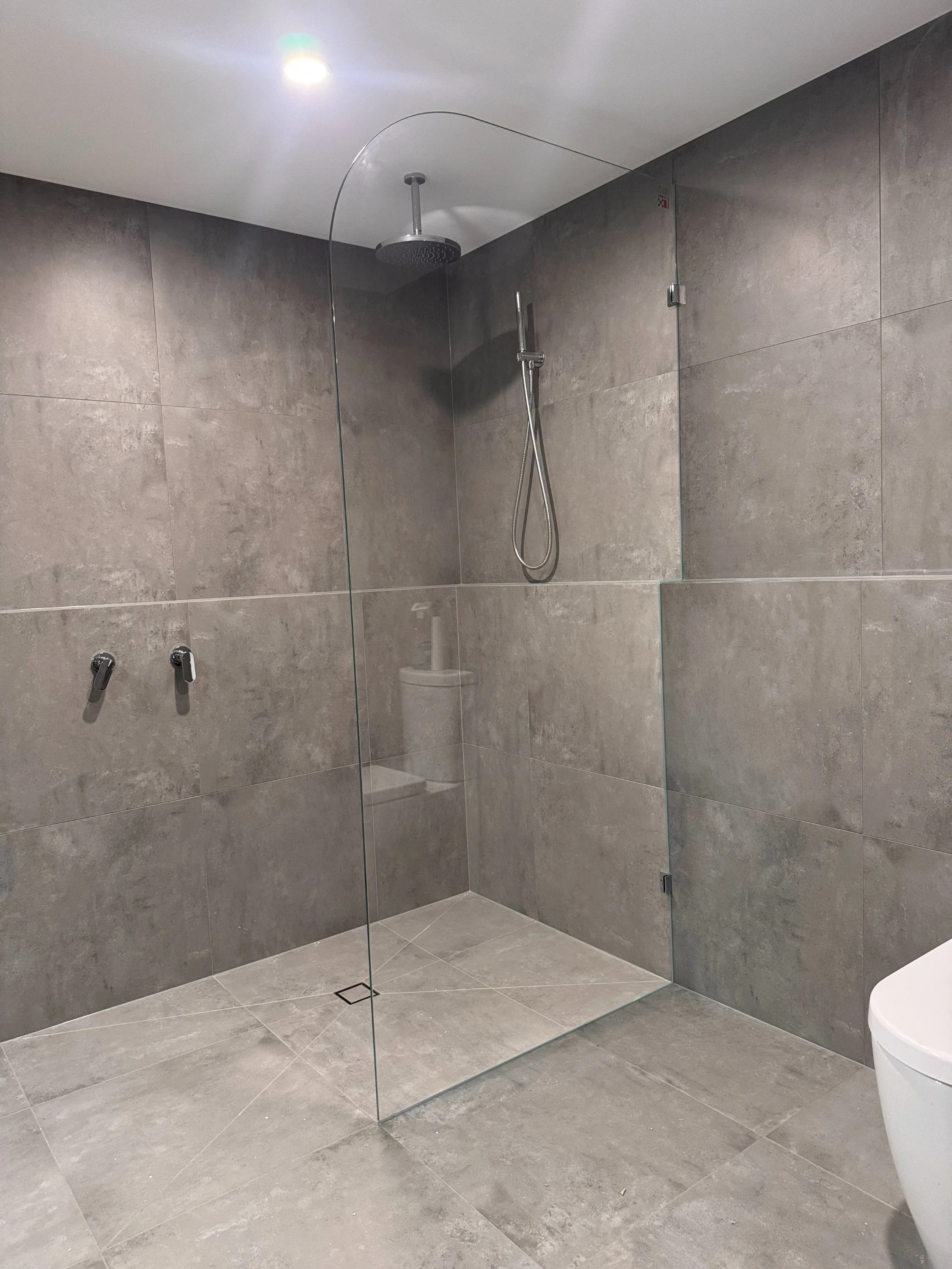 Modern walk-in shower with gray tiles, glass partition, rain showerhead, handheld shower, and a built-in bench, next to a white toilet.