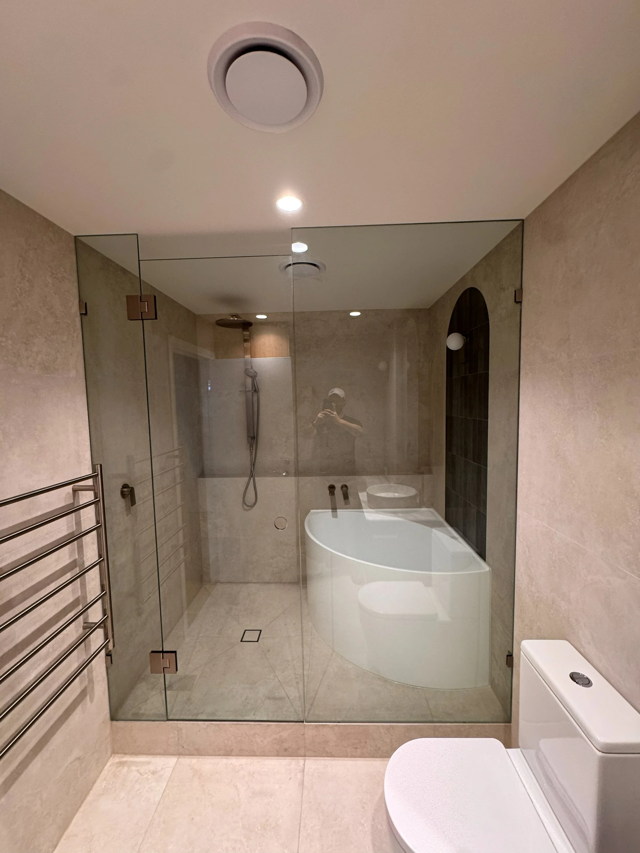 Modern bathroom with a glass-enclosed shower, walk-in shower with a handheld showerhead, bathtub, and a white toilet. The room features beige tile walls and floors, with recessed ceiling lights and a towel rack on the left.
