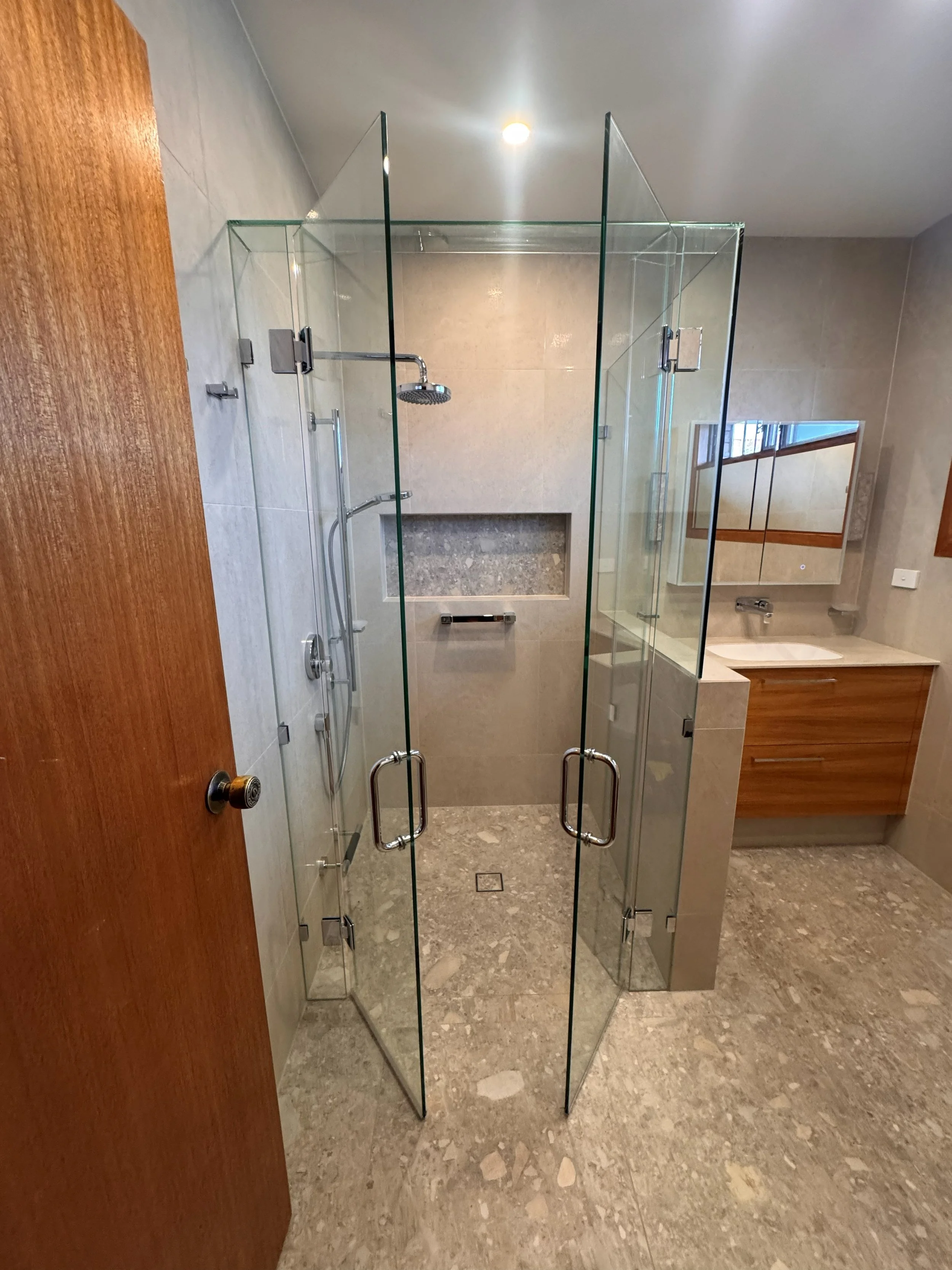 Modern bathroom with glass shower enclosure, wooden vanity with mirror, grey tiled walls, and beige marble floor.