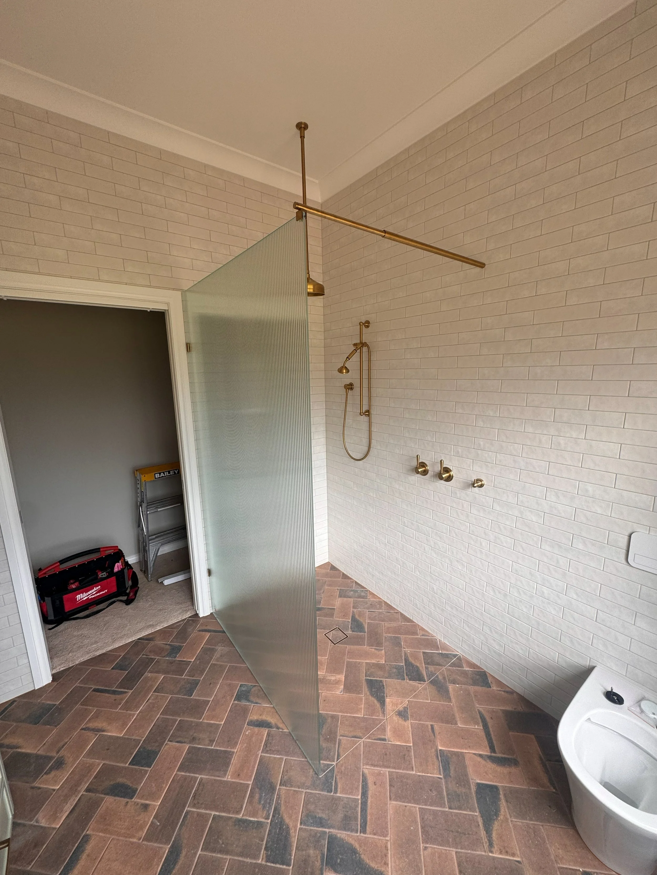 A modern bathroom with a brick-style tiled floor, a white wall with gold fixtures, a glass shower partition, and a door leading to a room with a toolbox and ladder.