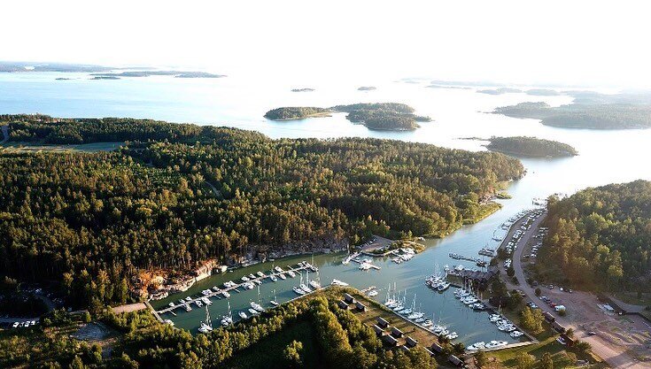 Ilmakuva venesatamasta metsäisellä alueella meren rannalla, useita veneitä laitureissa ja saaria taustalla auringonlaskun aikaan.