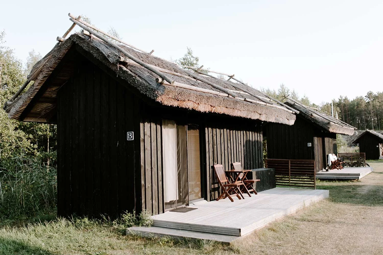 Puinen mökki ja terassi, jossa on kaksi tuolia ja pöytä. Mökissä on olkikatto ja se on numeroitu 13. Taustalla näkyy metsää.