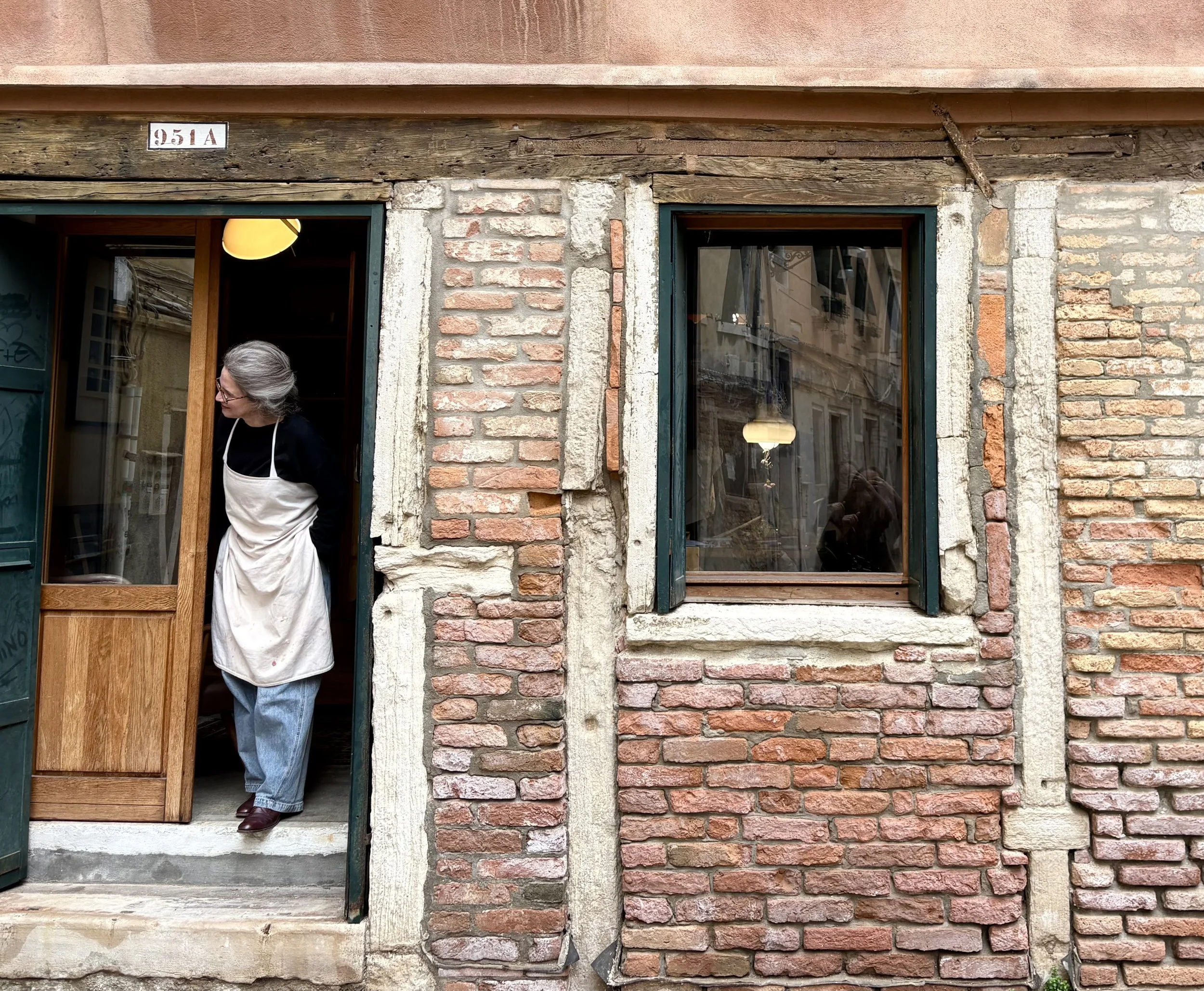 A woman with jeans, brown leather shoes, a long sleeved black top and white apron standing in the doorway of an old brick building with stope steps and wooden doors with glass panels