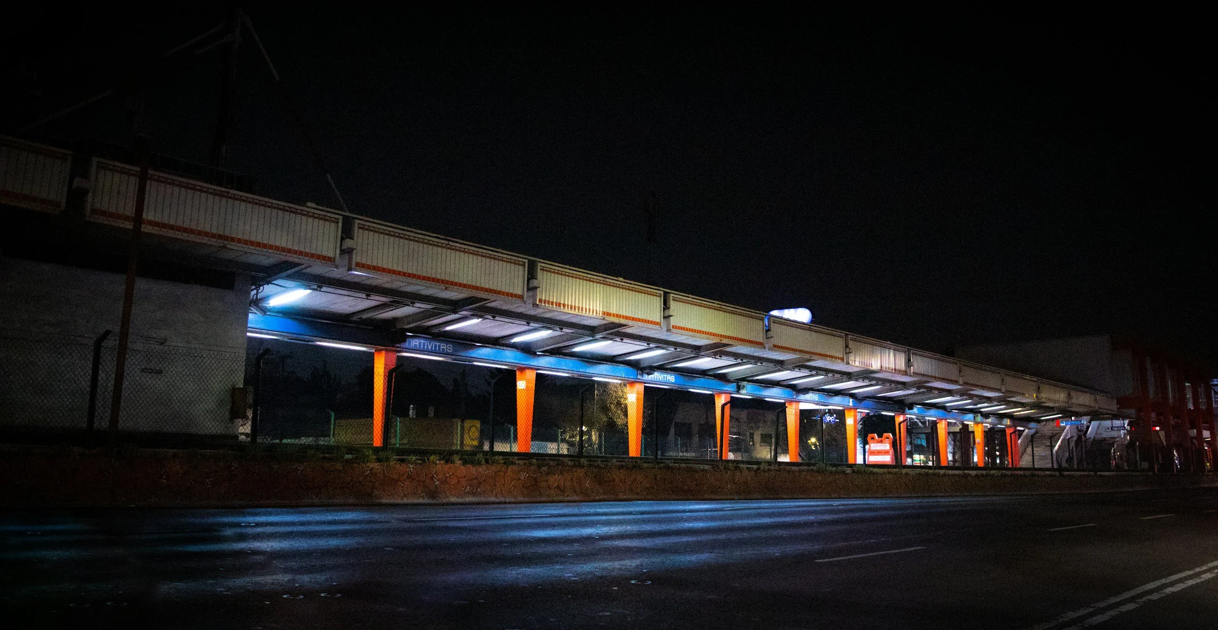Nighttime view of a bus stop shelter with lights, orange and blue accents, and a sign, located along a wet street with a fence on one side.