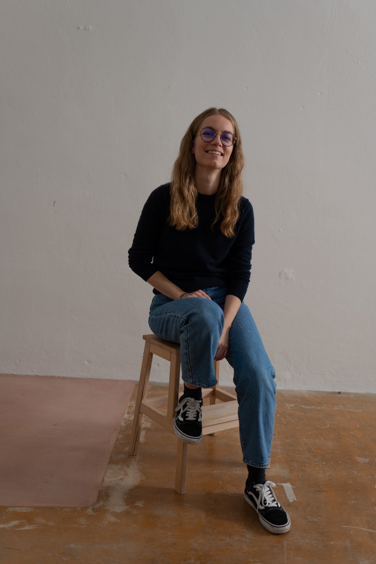 Junge Frau mit Brille, lockigem Haar sitzt auf einem Holzstuhl vor einer weißen Wand, trägt schwarze Schuhe, Jeans und schwarzen Pullover.