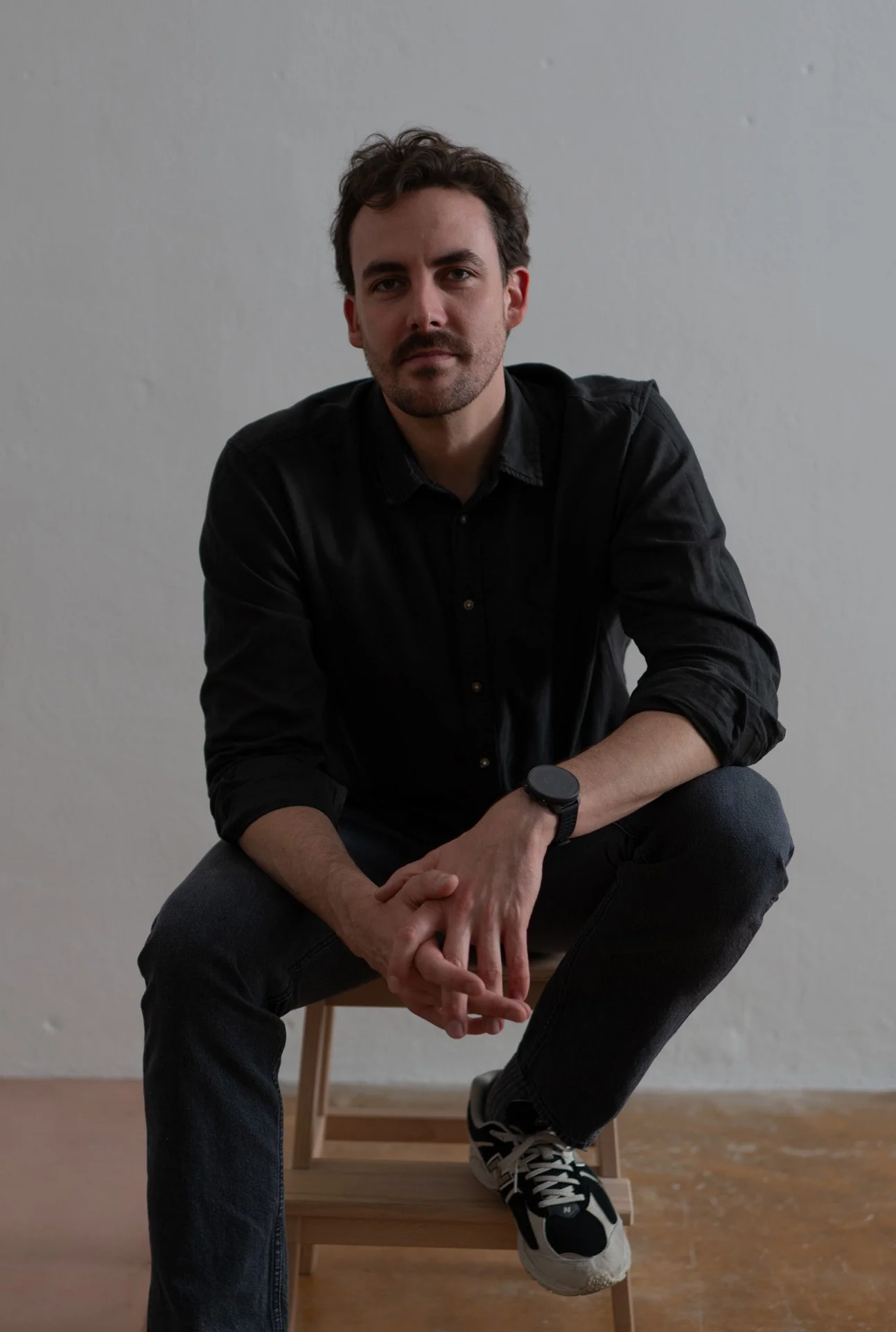 Ein junger Mann mit dunklem Haar, Bart und schwarzen T-Shirt sitzt auf einem Stuhl vor einer weißen Wand, Blick direkt in die Kamera gerichtet.