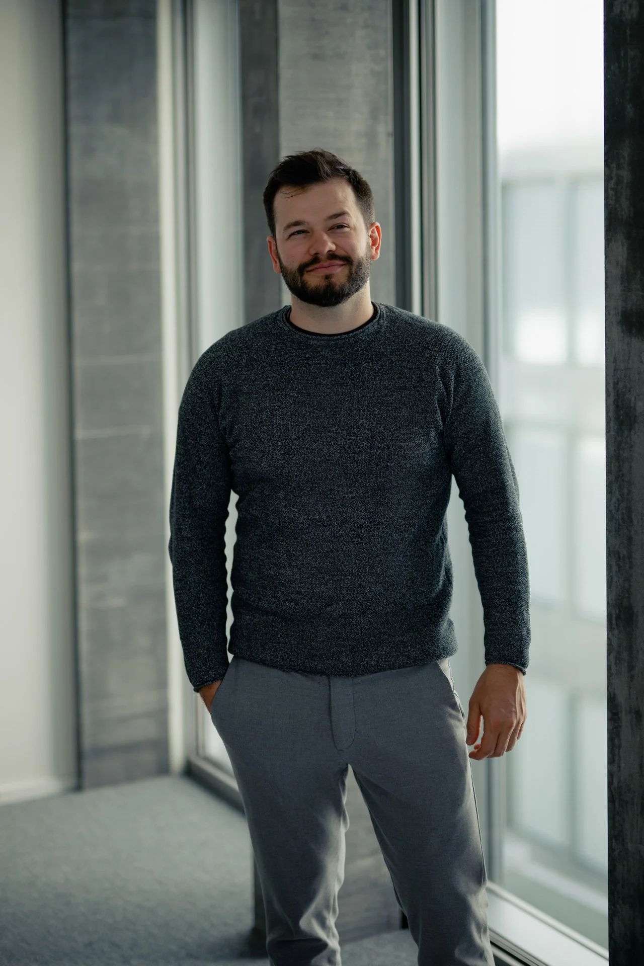 Ein Mann mit dunklem Haar und Bart steht in einem modernen Gebäude neben großen Fenstern.