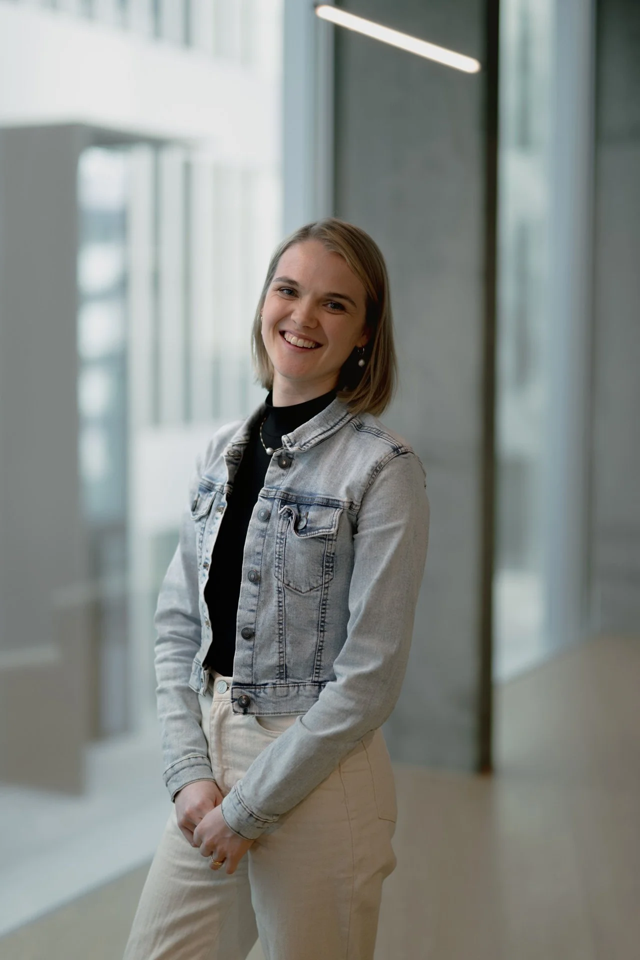 Junge Frau mit braunen Haaren lächelt, trägt eine Denimjacke und beige Hosen, steht in einem modernen, hellen Raum.