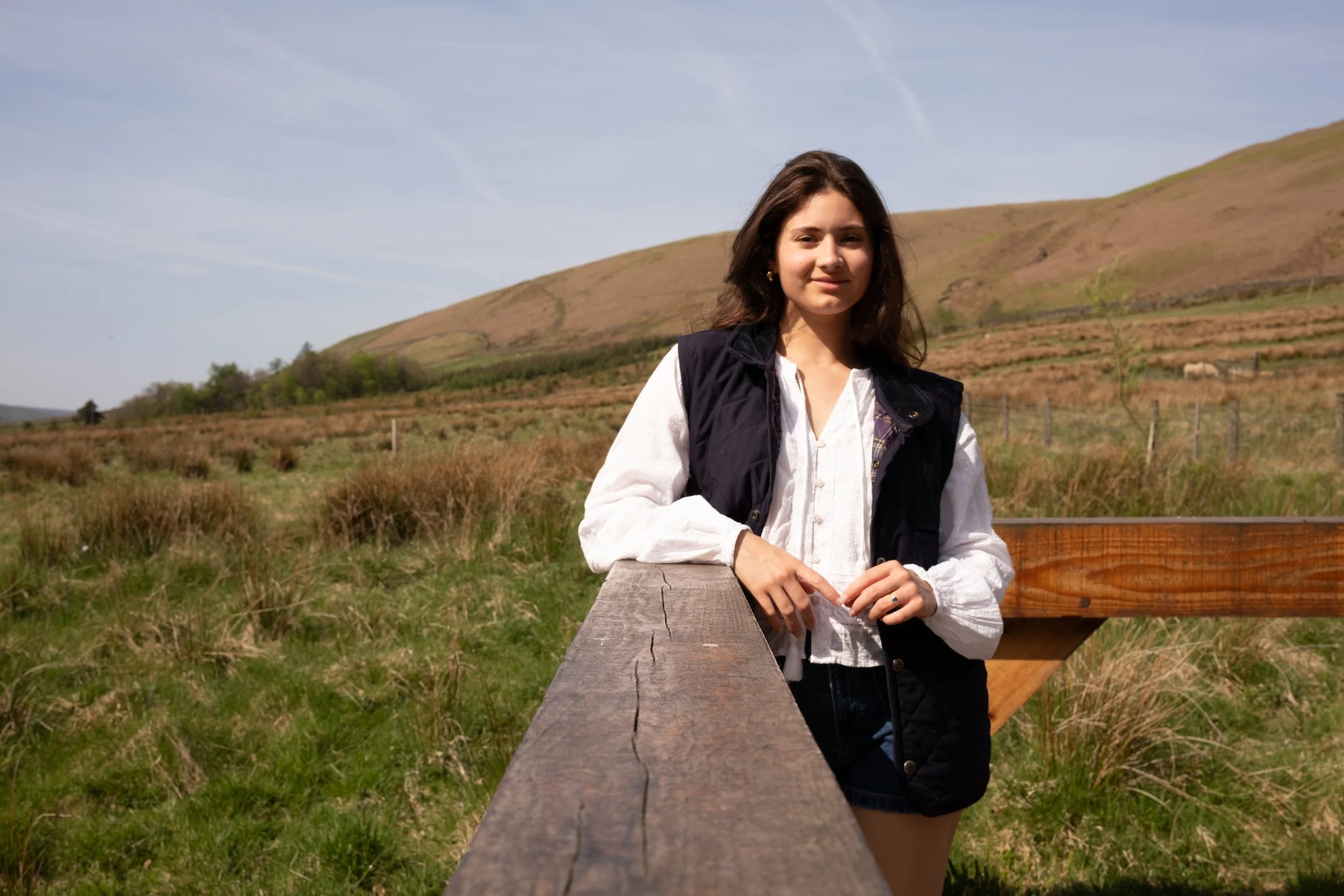 A young woman with brown hair leaning on a wooden railing outdoors in a rural landscape with rolling hills and grassy fields.