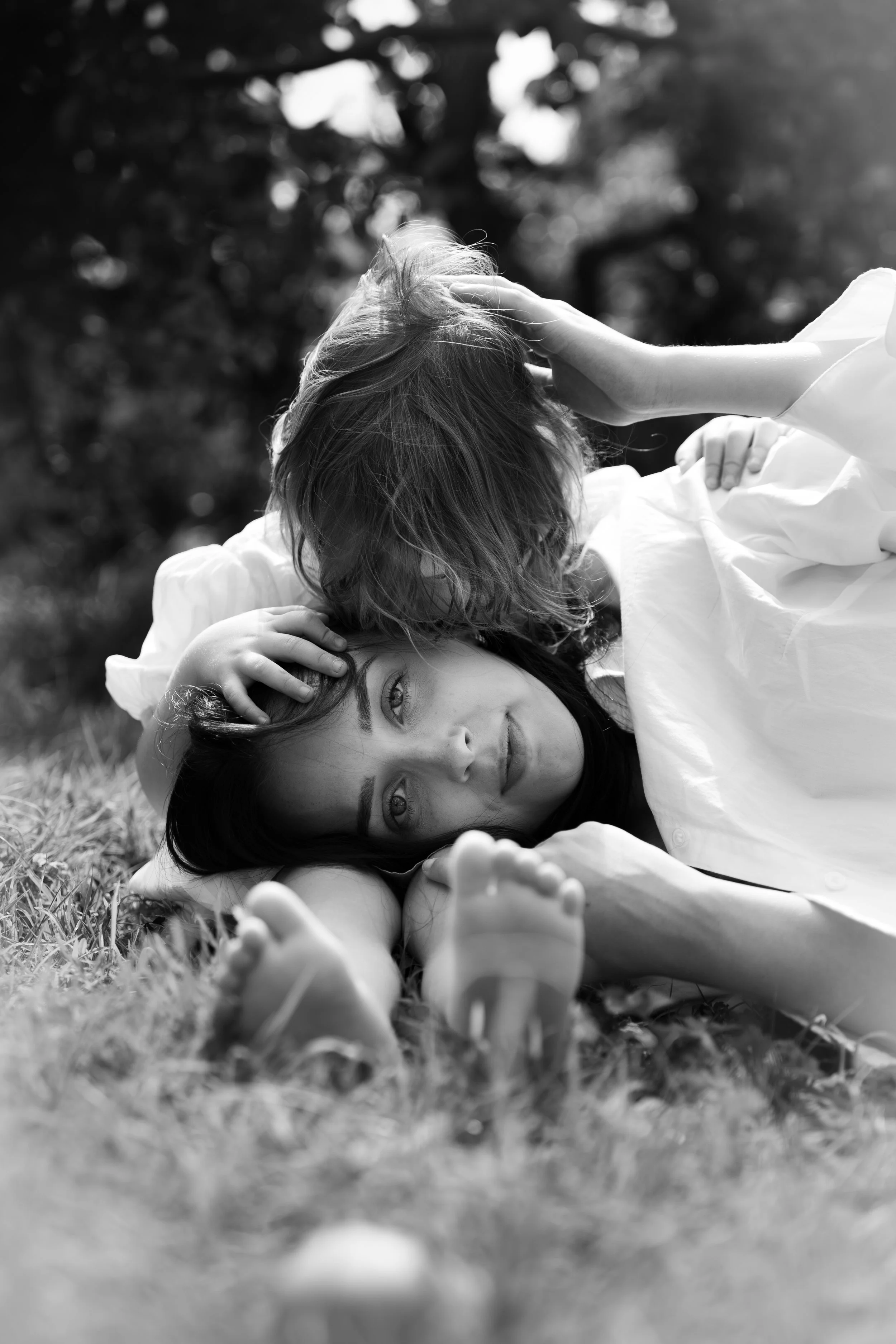 A woman lying on the grass outdoors, resting her head on the ground, with a child lying on her back. They are surrounded by nature, with a blurred background of trees. The photo is in black and white.