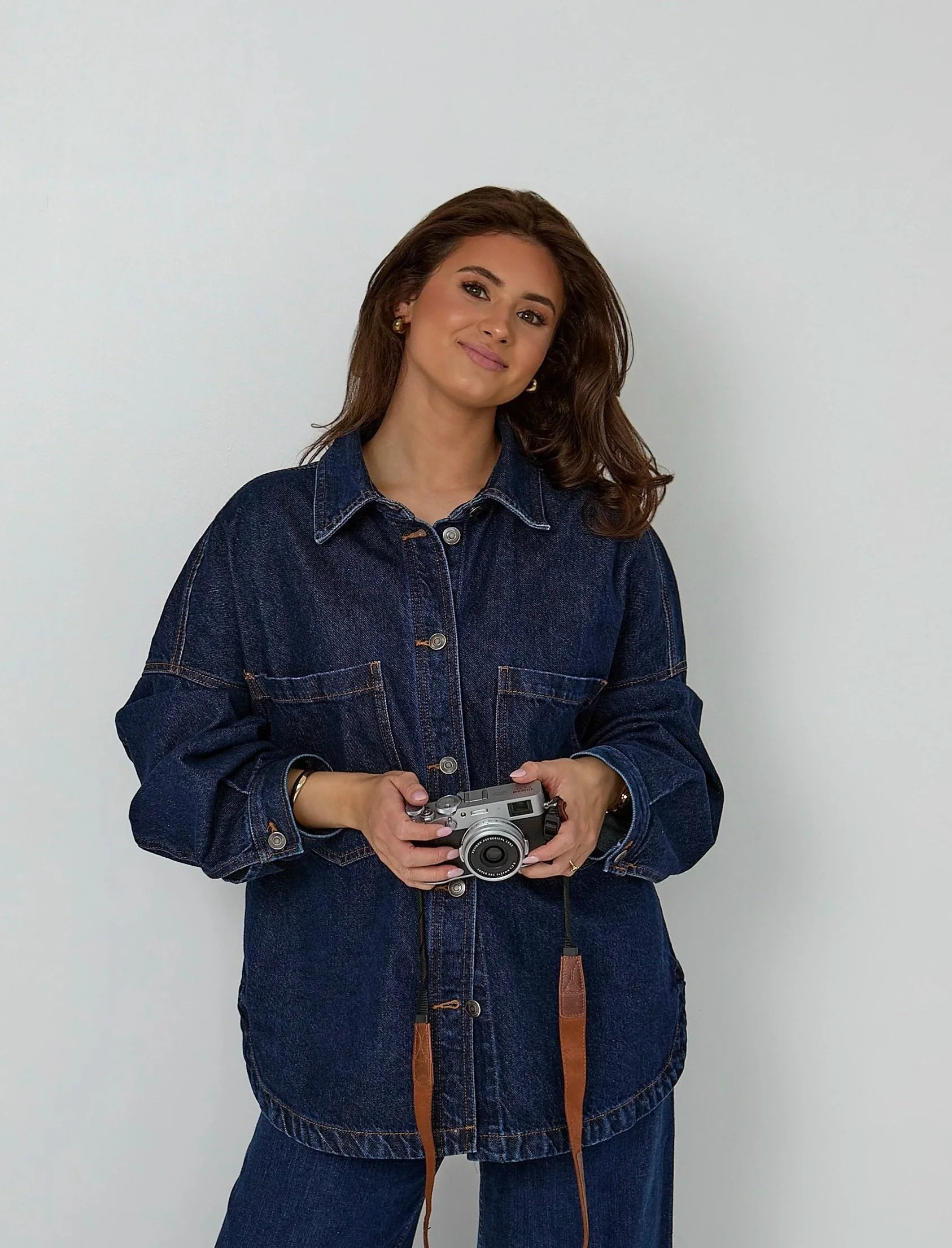 A woman with long, wavy brown hair and earrings holding a camera, wearing a dark blue denim jacket and jeans, standing against a plain light-colored wall.