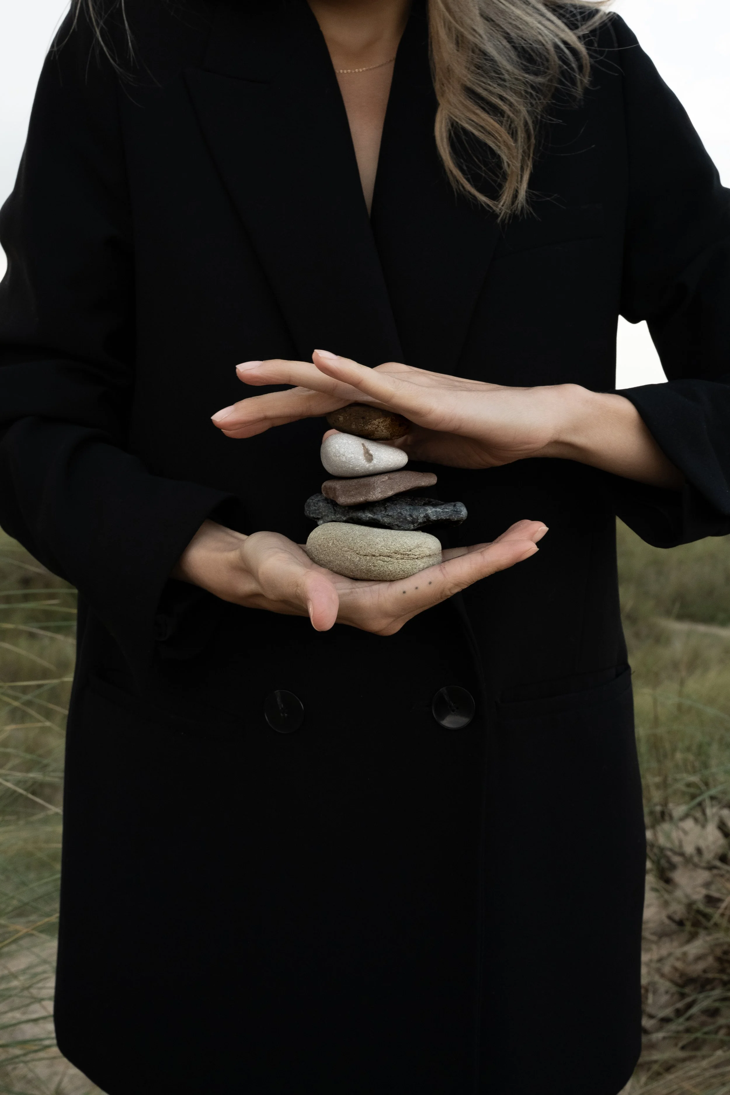 A woman in a black coat balances a stack of seven smooth stones outdoors.
