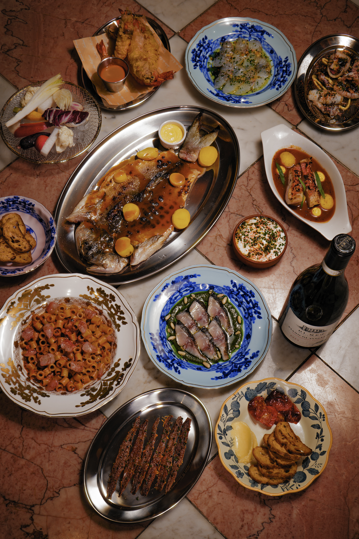 A variety of Italian dishes on a marble-tiled table, including a grilled fish with lemon slices, pasta with ham, sliced cured meats, raw fish carpaccio, fried vegetables, shrimp, grilled meat, and a bowl of tomato-based stew with vegetables, accompanied by a bottle of red wine.