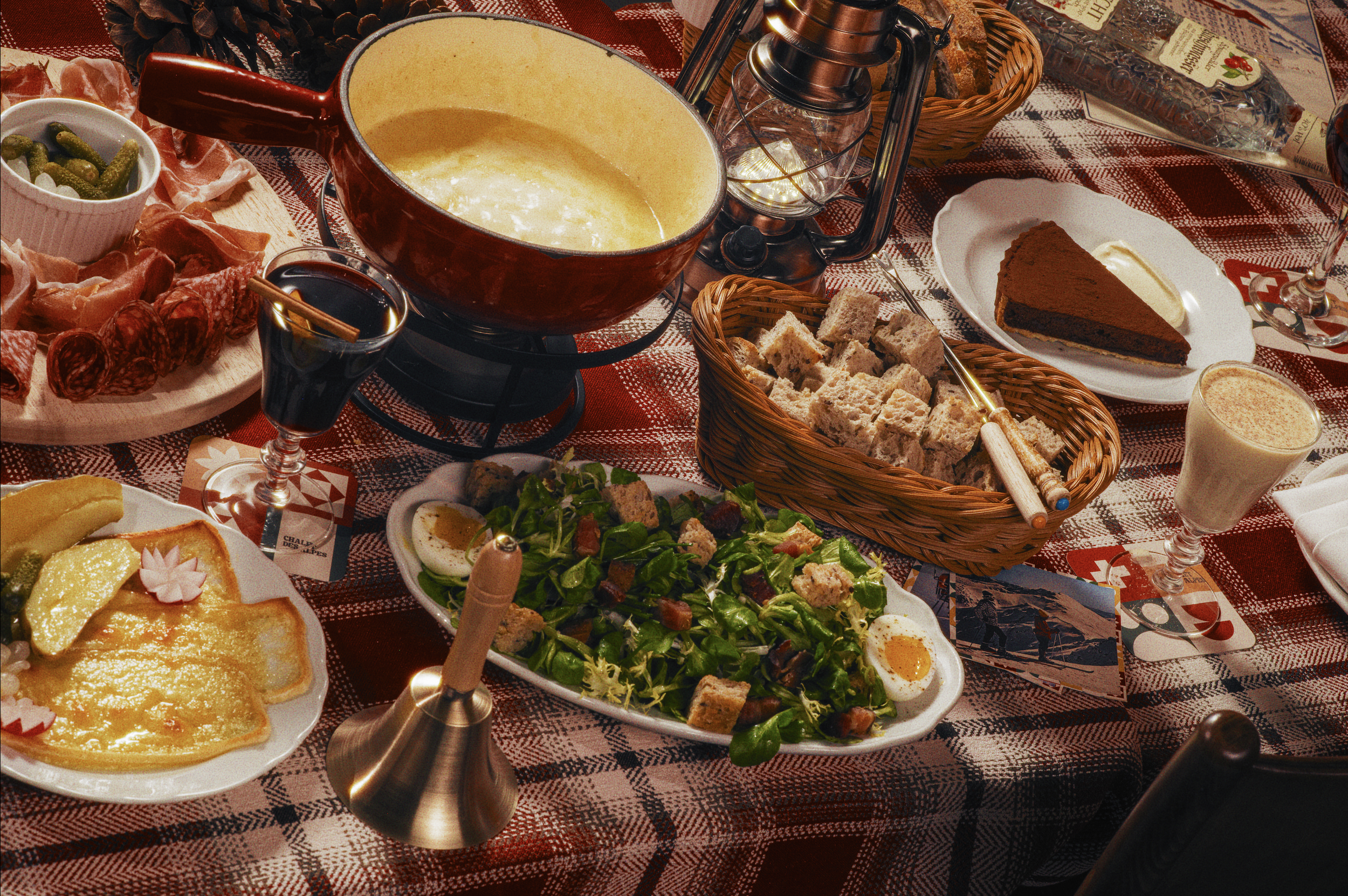 Table set for a meal with various dishes including a bowl of cheese, a salad, hard-boiled eggs, a meat platter, slice of cake, bread cubes, and a glass of red wine. The table is covered with a red checkered cloth and decorated with pinecones and holiday-themed items.