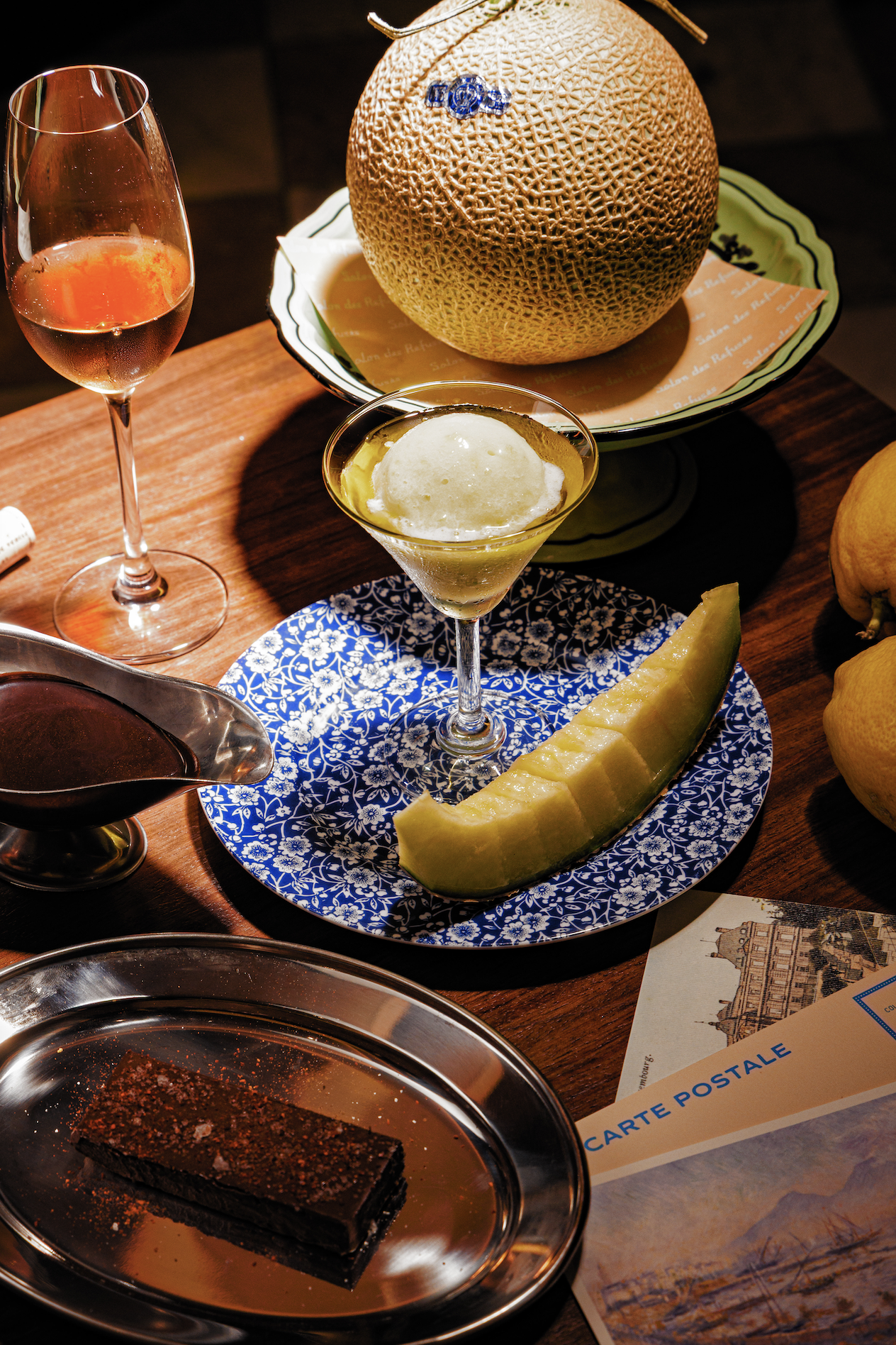 A cantaloupe melon on a decorative plate, a glass of rosé wine, a small scoop of gelato in a cocktail glass, a slice of honeydew melon on a blue patterned plate, and a piece of chocolate cake on a silver tray.