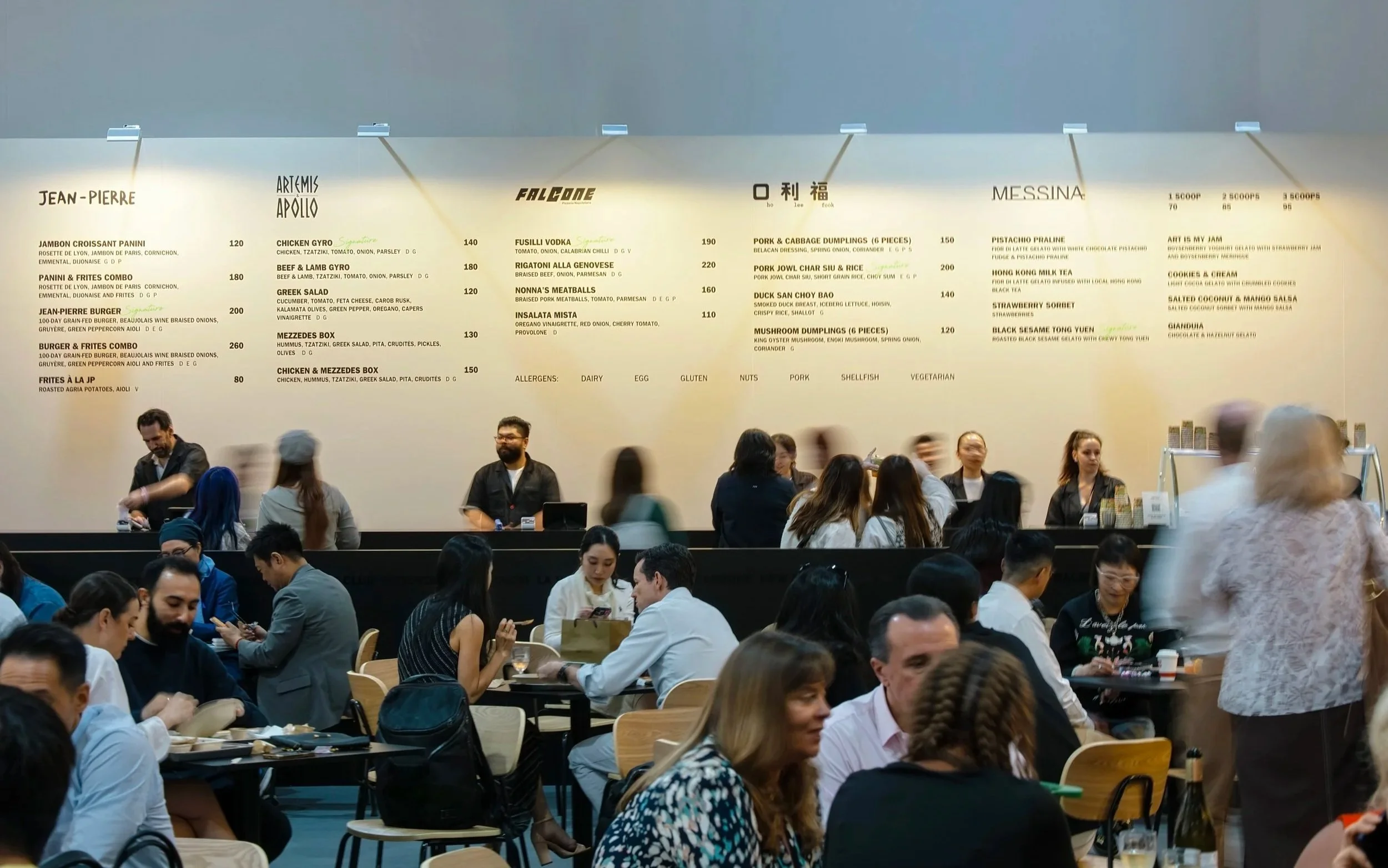 A busy restaurant with many patrons seated at tables, some looking at their phones, and staff behind the counter with a large menu board overhead displaying various dishes and prices.